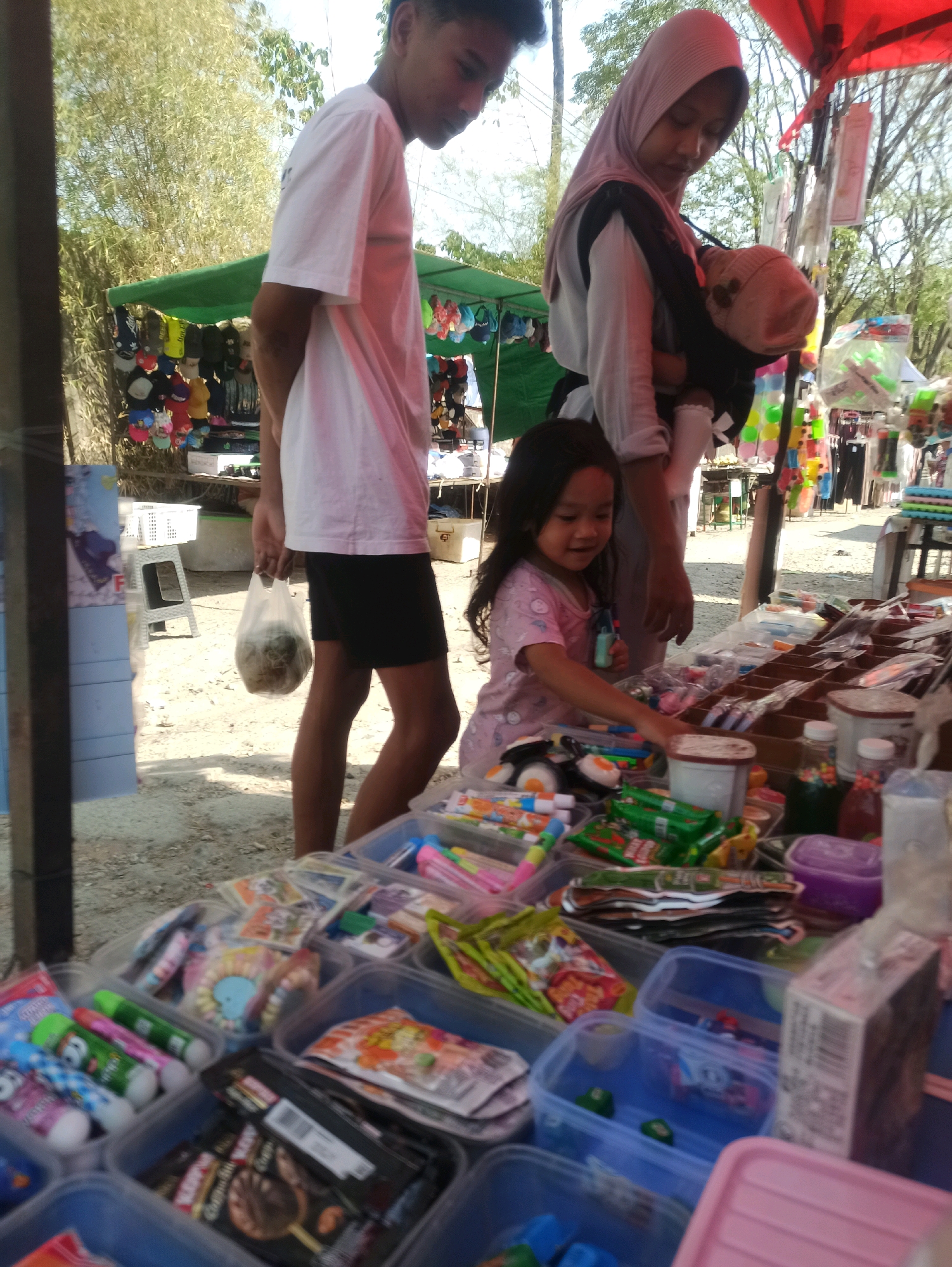 In Telagamurni, Indonesia, an outdoor market stall displays a variety of colorful goods. A young girl, wearing a pink shirt adorned with cartoon characters, actively browses the merchandise, her right hand reaching out towards the items on the table. Beside her, a woman in a pink hijab carries a baby in a carrier, looking down at the stall. To the left, a young man in a white t-shirt and black shorts holds a plastic bag filled with what appears to be food items. The stall is filled with an assortment of candies, snacks, and packaged goods, arranged in clear plastic bins. The ground is covered in dirt and gravel, with a white plastic chair visible behind the man. The background features stalls with various items, including hats and clothing, and a backdrop of lush green trees and foliage, suggesting a bright, sunny day.