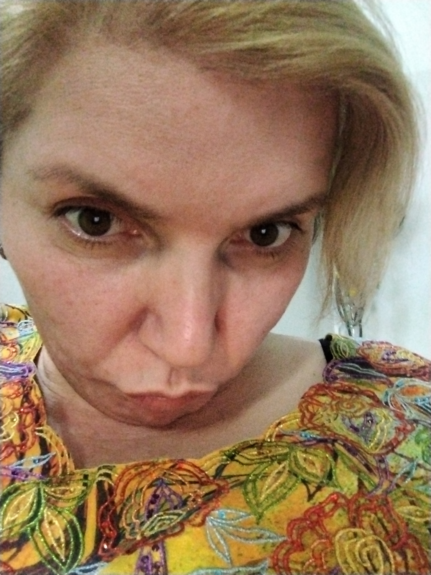 A close-up selfie shows a fair-skinned woman with short, blonde hair and dark brown eyes. Her expression is somewhat pouting. She wears a bright yellow top with an intricate floral pattern embellished with sequins or beads in various colors like green, red, orange, and purple. The background is plain and appears to be a white wall. There's no indication of the location being Al Mas'oudi, United Arab Emirates, and no information indicates a connection to that city.