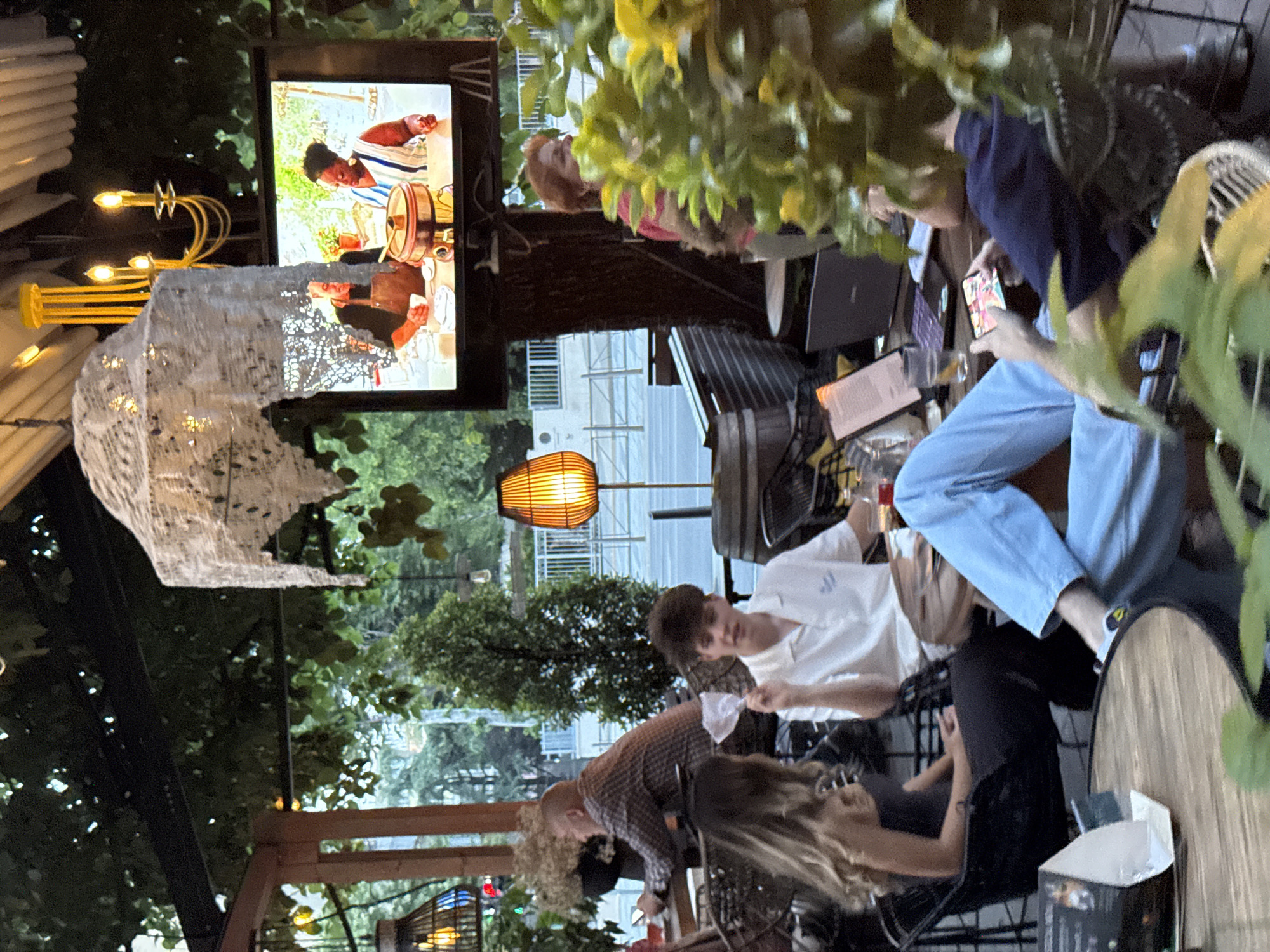 People are sitting at an outdoor patio in Belgrade, Serbia. There are five people at a table, some using electronic devices. There is a television screen showing a cooking program. Overhead is a chandelier and decorative lighting. Trees are visible.
