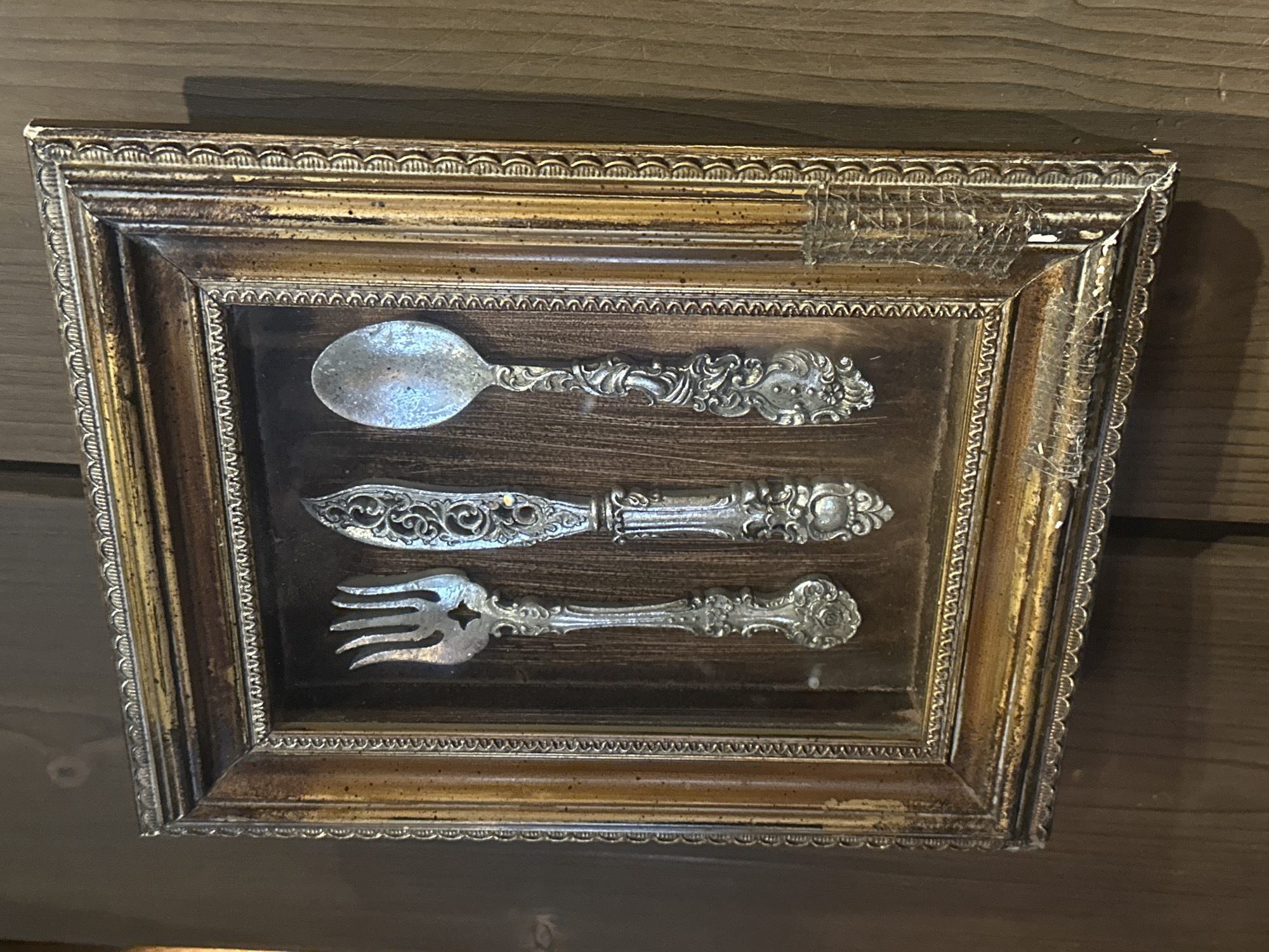 A framed display of antique silverware is mounted on a wooden wall. The frame is ornate, gold and silver, with some damage and what appears to be cobwebs in the upper right corner. Inside the frame, a spoon, knife, and fork are arranged vertically against a dark wood background. The silverware pieces are ornate and made of a light-colored metal, possibly silver. The location is unknown.
