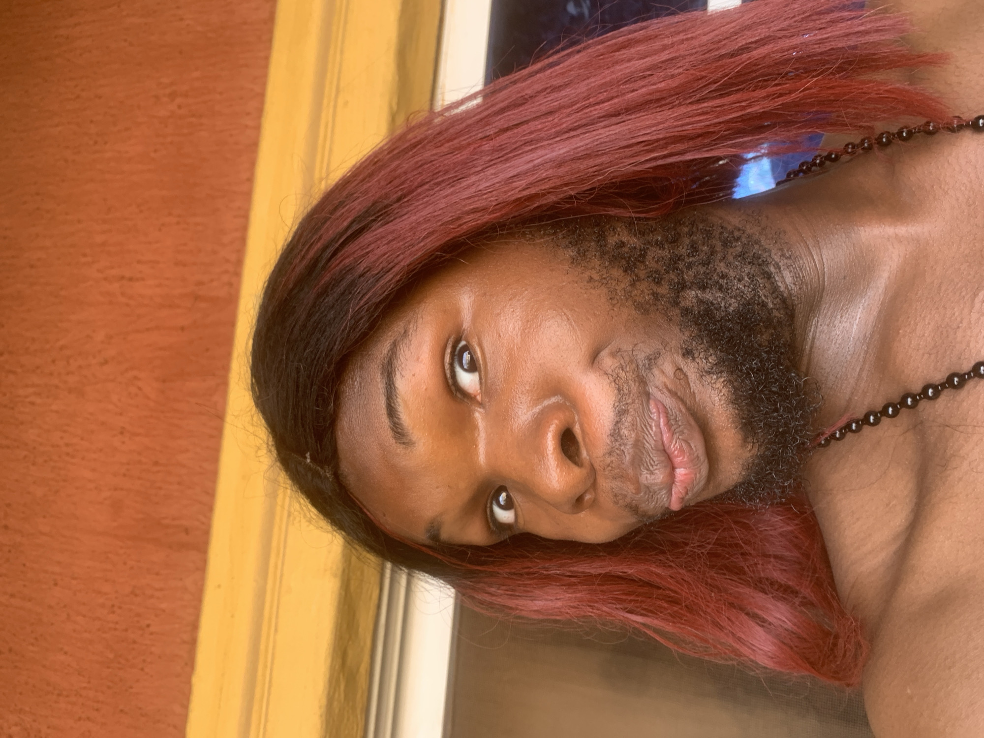 This close-up portrait features an individual with dark skin, prominent dark facial hair (beard/stubble), and long, reddish-brown hair. The image is rotated 90 degrees clockwise, showing the person's head tilted, with their eyes looking directly at the camera. A neutral to slightly contemplative expression is evident, accompanied by a subtle smirk or soft smile. The person wears a dark beaded necklace around their neck.
The background is composed of a textured, reddish-orange wall to the left, and a bright yellow vertical architectural element, possibly a doorframe or pillar, to the right. A blurry area behind the subject suggests a reflective surface or window, displaying hints of blue and white light. The bright, natural-looking illumination indicates a daytime setting. This candid moment is captured in Nkpor, Nigeria. There is no visible text in the image.