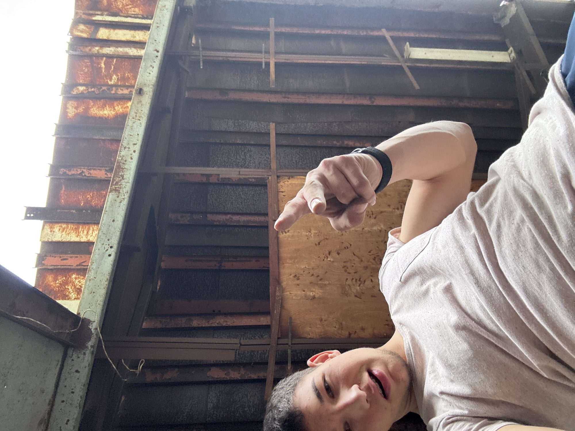 An upward-facing photograph captures a young man in the foreground pointing towards the weathered ceiling or underside of a structure in Zlatograd, Bulgaria.
The main subject, a young man with short, dark hair, is visible from the chest up in the lower right portion of the frame. He is wearing a light-colored t-shirt and a dark wristband on his left arm. His mouth is slightly open, and his eyes appear to follow the direction of his right index finger, which is extended and pointing towards the upper center of the image. His expression is neutral.
The background reveals the complex internal structure of an old building, likely a roof or elevated platform. It is characterized by a mix of materials: dark, aged wooden planks and beams form a grid-like pattern, while rusted corrugated metal sheets are prominent, especially towards the brighter left side. Several metal beams, some heavily corroded with a greenish-white patina, crisscross the scene. Bright daylight streams in from the upper left, illuminating the rusted surfaces and suggesting an opening to the outside. Some thin, light-colored wires or string hang loosely from the structure. The overall impression is one of an old, possibly abandoned or disused industrial, agricultural, or storage building, showing significant wear and signs of decay.
The activity depicted is the man drawing attention to something within the overhead structure by pointing. The time of day is clearly daytime due to the bright natural light. There is no visible text in the image.