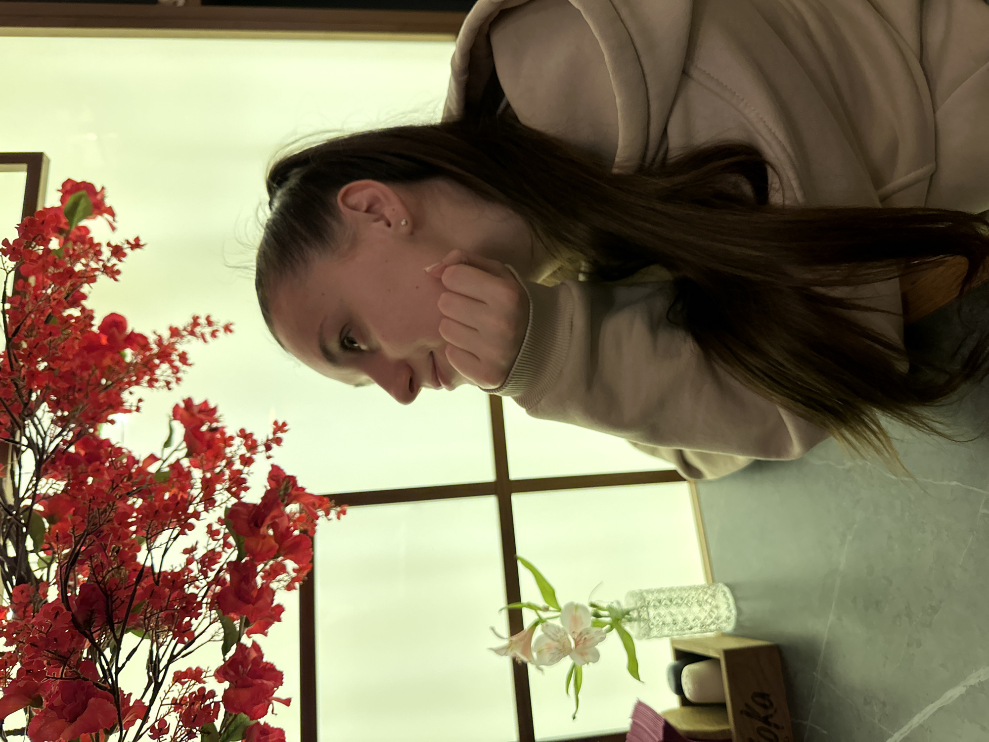 A woman with dark hair pulled back in a ponytail is seated at a table indoors, near a window. She is wearing a beige hooded sweatshirt and resting her chin on her right hand. To her left, there is a vase with red flowers. In front of her, there is another vase with white flowers. Below the second vase, there is a wooden stand, with "Soko" printed on the front of it. The location is Rostov-on-Don, Russia.