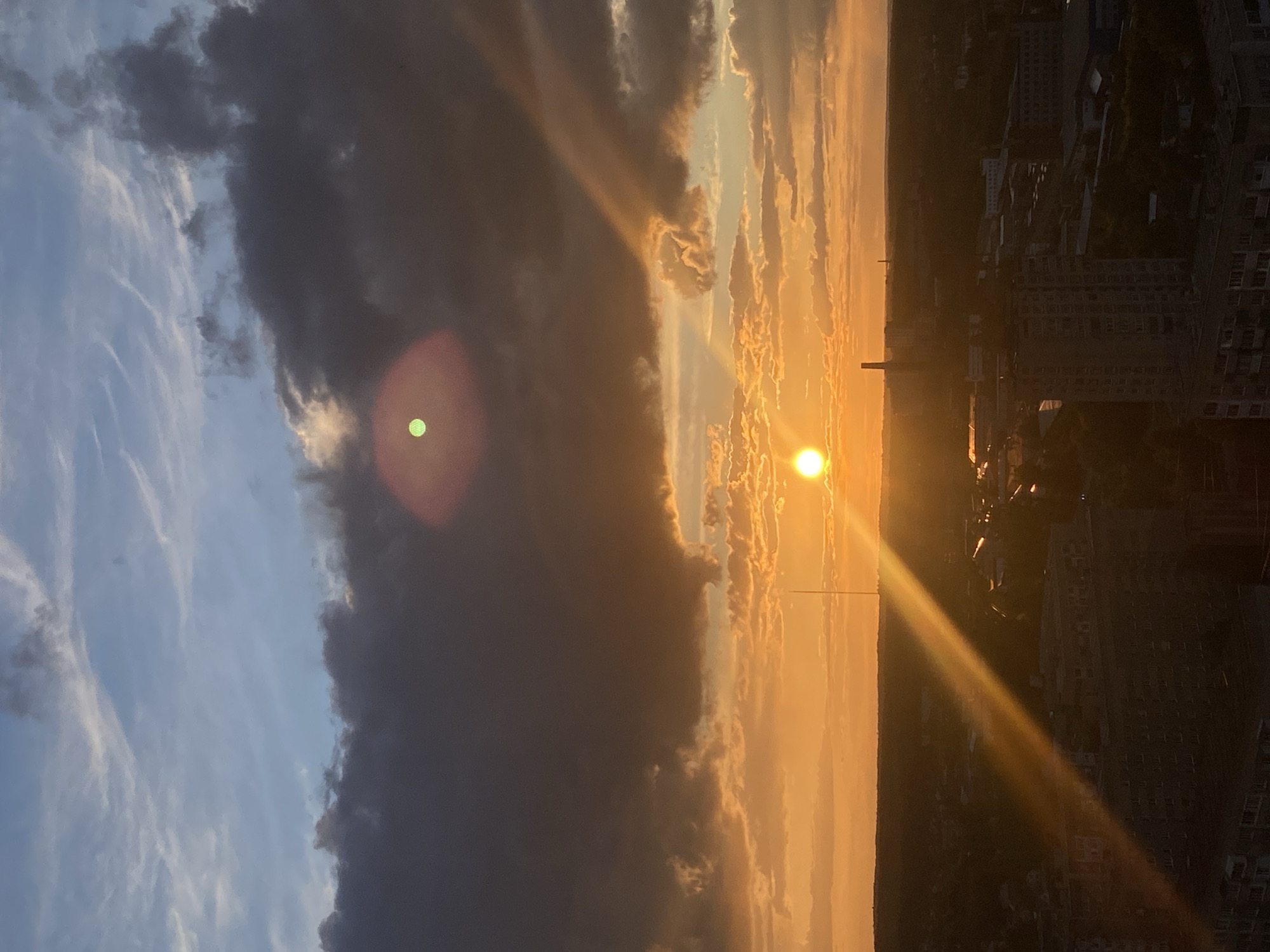 The image is a rotated view of a sunset in Izhevsk, Russia, as suggested by the urban landscape silhouette on the right side. The sky is divided between a light blue area with thin, white clouds on the left and a massive, dark cloud formation dominating the center. The sun, appearing as a bright orb, is near the horizon on the right. Sunlight emits rays across the scene, illuminating the edges of the clouds and casting a warm glow over the landscape. The city skyline is visible as a dark, textured area, with building silhouettes and possible infrastructure structures. The lens flares are visible in the center and left portions of the image. The angle seems to be captured from a high vantage point.