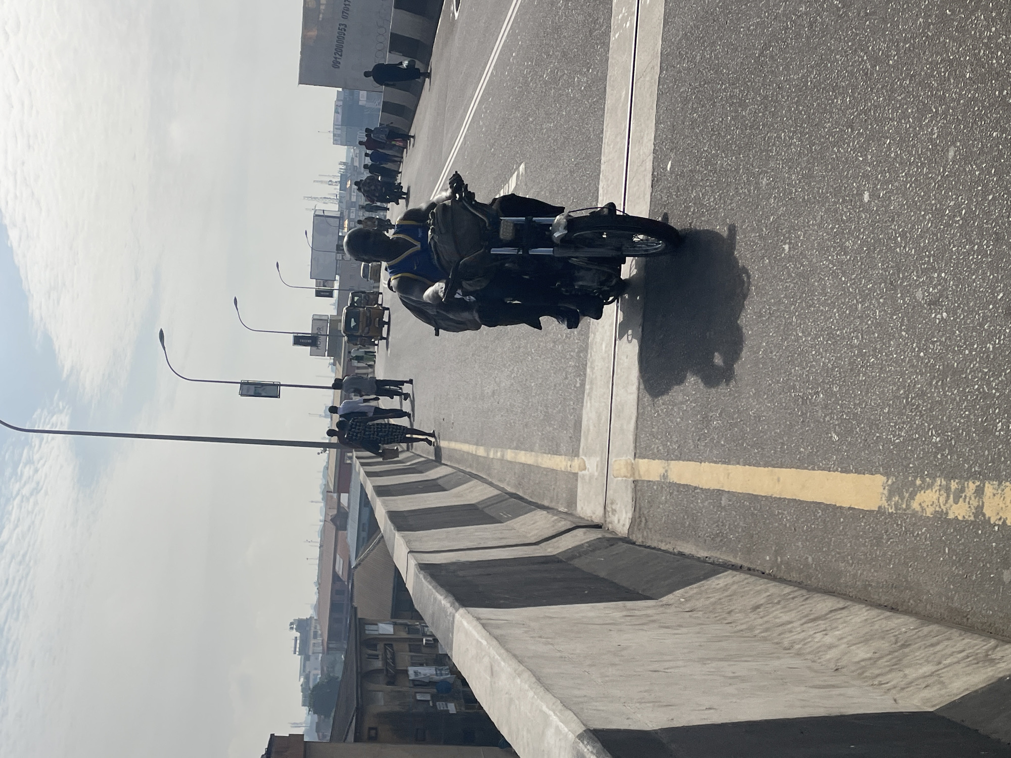 The image depicts a scene on a bridge in Ikeja, Nigeria. A motorcycle with two riders is moving along the road. Pedestrians are walking on the side of the bridge. Streetlights line the road. Buildings are visible below the bridge. A painted yellow line marks the edge of the road.