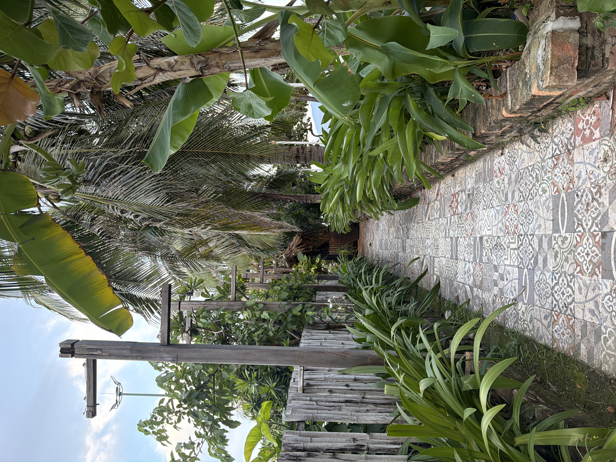 The image depicts a pathway in Nha Trang Ward, Vietnam. The pathway is made of patterned tiles and is bordered by lush green foliage on both sides. On the left, a rustic fence made of bamboo and wooden posts separates the pathway from more greenery and a tall lamppost in the distance. On the right, the pathway is lined with a low brick wall and more tropical plants, including banana trees and palm trees, which create a dense canopy overhead. The scene appears to be an outdoor area, possibly a garden or the grounds of a resort, with a hint of a thatched roof structure visible in the background. The sky is partly cloudy.