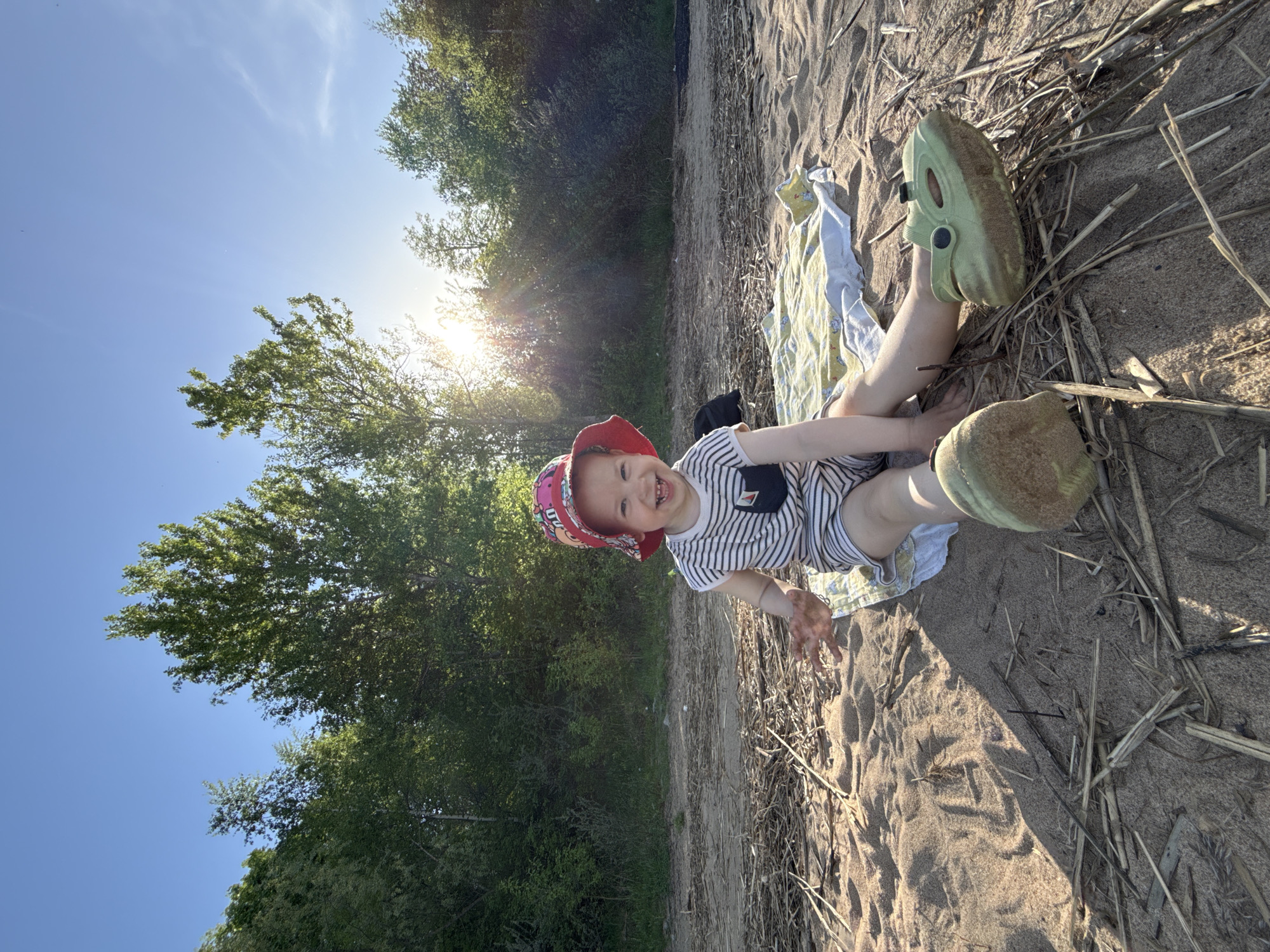 A young child wearing a red and pink hat, a striped shirt with a small pocket, shorts, and olive-green Crocs is lying on a beach in Шебунино, Russia. The child is on a blanket patterned with animals. The beach sand is mixed with dry sticks. The background shows trees and a blue sky with the sun shining through the leaves.