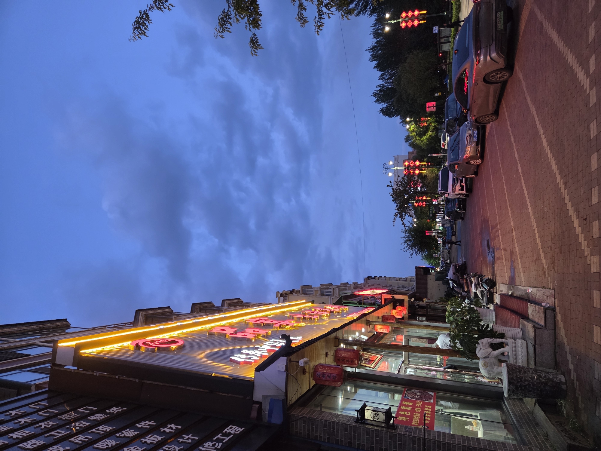 This media file captures a commercial street scene in Tongjiang, China, during dusk or early night. The image is rotated 90 degrees clockwise, showing a street lined with multi-story buildings under a dark blue, cloudy sky.
The prominent feature is a building on the left, illuminated by a large, bright yellow-orange neon or LED sign displaying Chinese characters. Below this main sign, other signs with Chinese text advertise services such as "暖管件" (heating pipe fittings), "金板材" (metal sheets), and "工" (work/construction). Red Chinese lanterns hang from the building's facade, adding to the cultural ambiance. Near the entrance, a small red sign advertises "真材实料老汤面,添加,干净卫生" (Authentic old broth noodles, fresh additions, clean and hygienic). A white stone sculpture, possibly an elephant, stands by the entrance, and a person is faintly visible inside the storefront.
On the right, a brick-paved street is visible, with several cars parked parallel to the curb. License plates like "OLB063" and "000683-08" are discernible on two of the vehicles. Further down the street, multiple scooters and motorbikes are parked on the sidewalk. Streetlights with red lantern-like decorations illuminate the road in the distance. Trees line the street, their dark foliage silhouetted against the twilight sky. The overall atmosphere is that of a bustling commercial area winding down for the evening, with businesses still active and lit up.