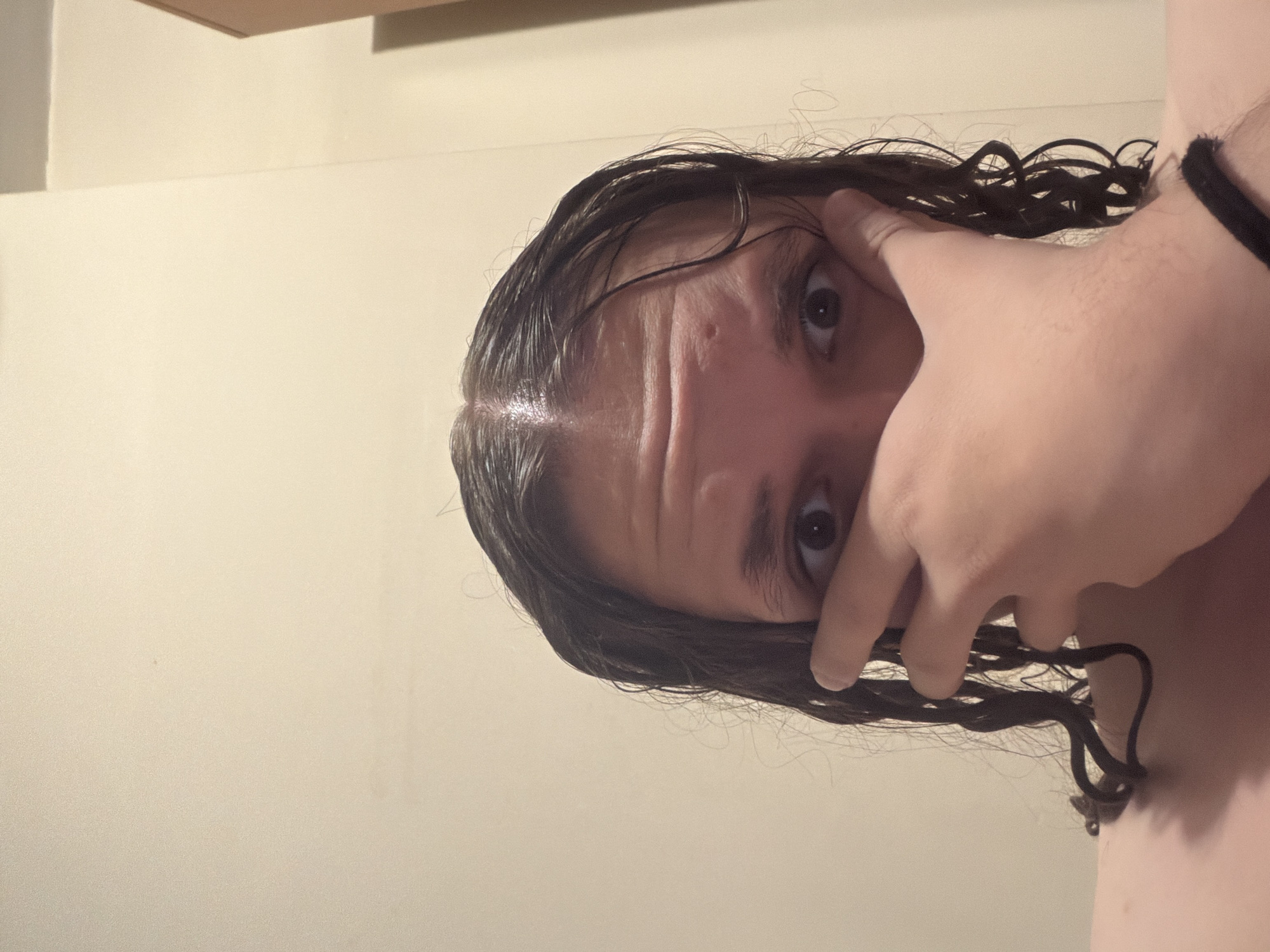 A medium close-up shot features a person with long, dark, wet or oily hair, taken indoors against a plain, light-colored wall. The image is oriented sideways, showing the person's head tilted with their left eye gazing directly towards the viewer. Their right hand is positioned over the lower right side of their face, partially obscuring it, with fingers gently spread. The visible left eye is wide open, and there are distinct horizontal lines or wrinkles on the forehead. A small, dark bump is noticeable on the left temple area. The dark, wavy or curly hair appears damp, clinging to the forehead and falling around the face, with some strands visibly caught between the fingers of the covering hand. A black hair tie or band is worn on the person's right wrist. The background is a uniform, light beige wall, with a subtle architectural feature or shadow visible in the upper left portion of the frame. The lighting is soft and appears to be artificial. No text is visible in the image. The general location is Plovdiv, Bulgaria, though no specific cues to this are present within the visual content.