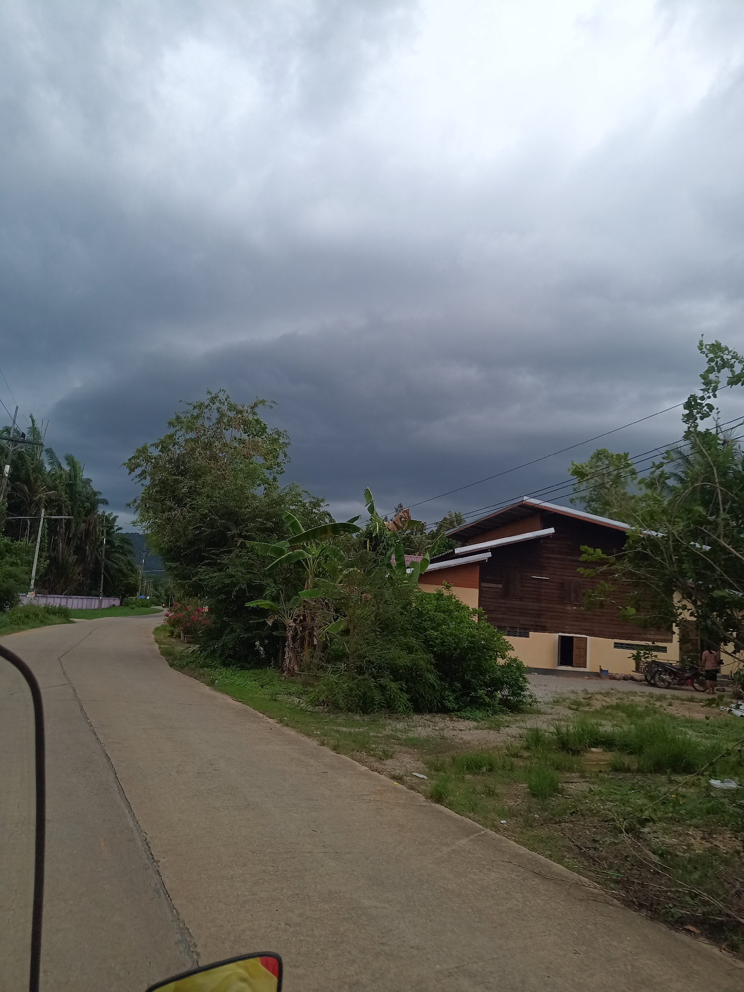 The photograph shows a paved road curving to the left in the city of Pak Phraek, Thailand. The sky is overcast with dark storm clouds. Lush greenery borders the road on both sides. A wooden building with a beige base is visible to the right, near the bend in the road. A motorcycle is parked near the building, where an individual stands. Power lines run overhead.