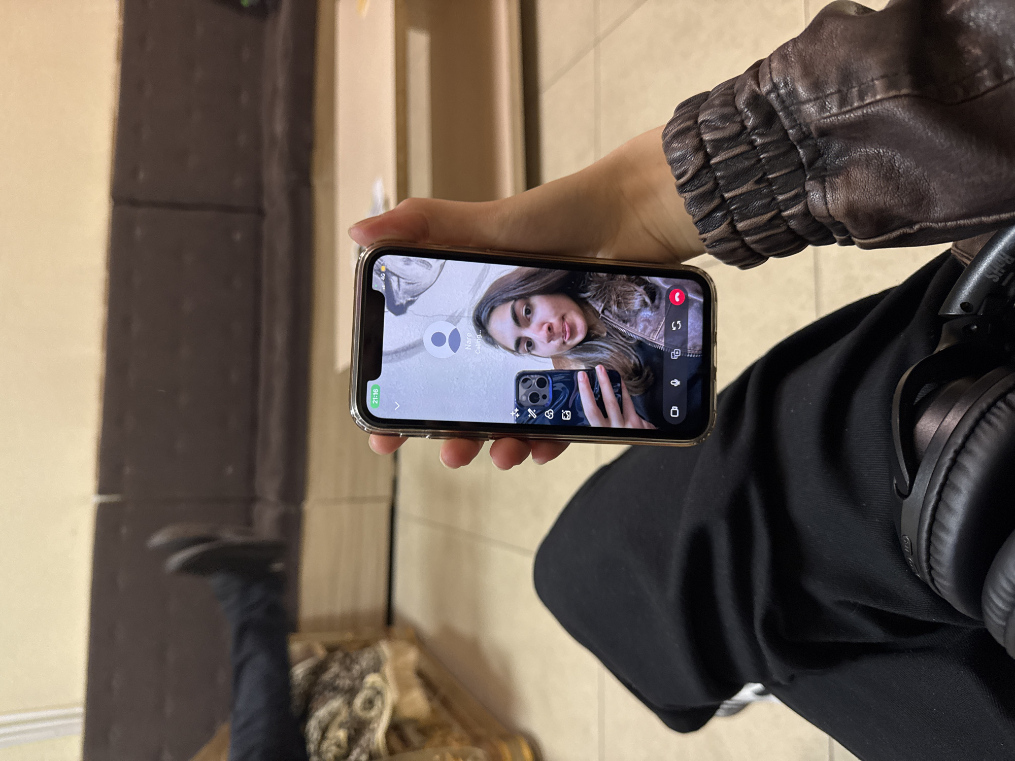 A person wearing a brown leather jacket and black pants is holding an iPhone in their hand. The iPhone is displaying a video call with another person. The background features a brown and beige wall, and a portion of a patterned object on the floor. Headphones are also visible next to the black pants. It is not possible to determine the exact location in Yerevan, Armenia from this image alone.