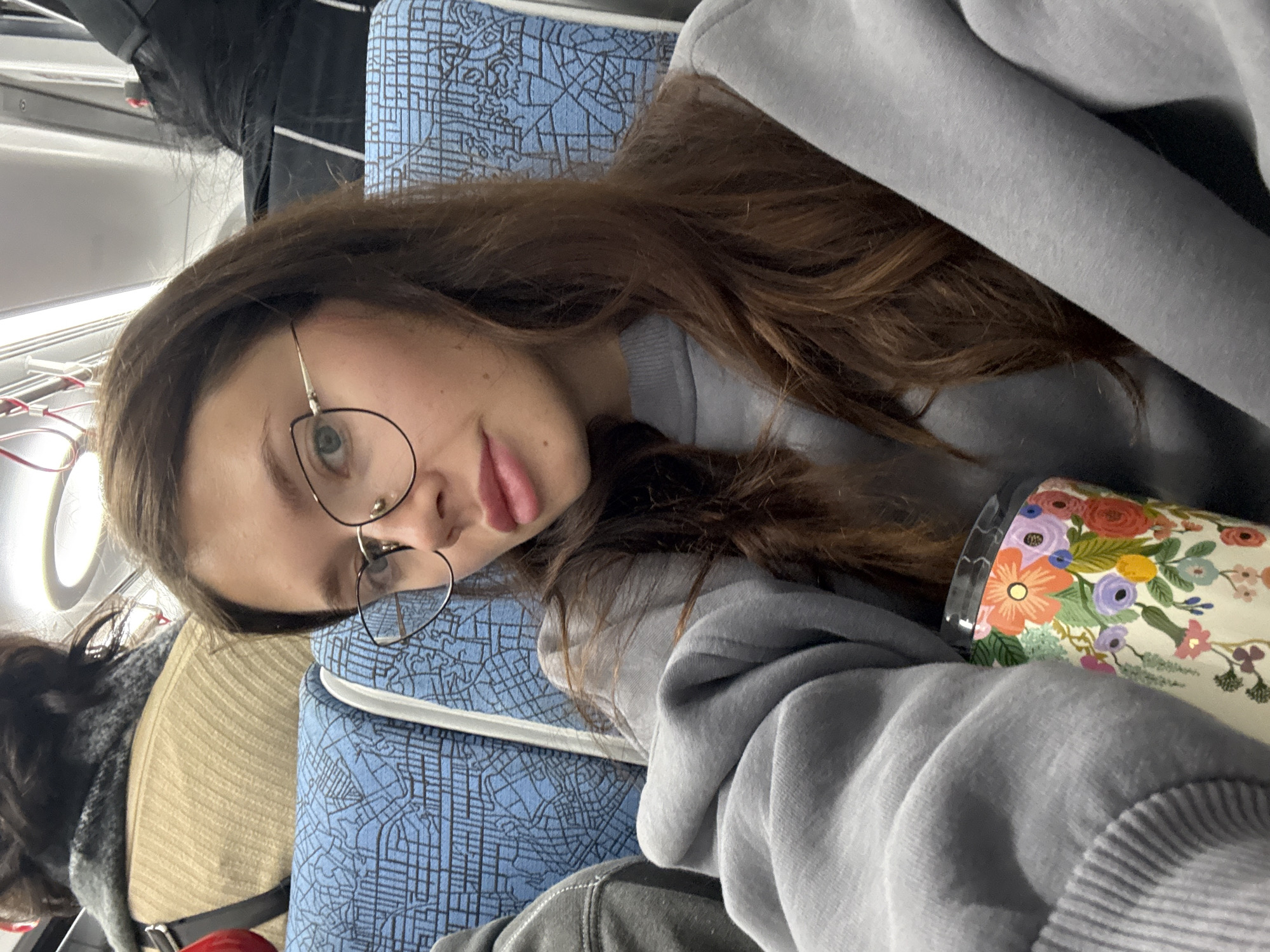 A young woman is featured in a close-up, selfie-style photograph taken inside what appears to be a public transport vehicle, such as a train or bus, likely in Tel-Aviv, Israel. She has long, wavy brown hair, wears round, thin-rimmed glasses, and is dressed in a light grey hooded sweatshirt. Her expression is casual and slightly pouting as she looks directly at the camera.
She is positioned against a seat covered in distinctive blue fabric patterned with a white, abstract grid or map-like design. To her right, a white travel mug or tumbler with a vibrant, multi-colored floral pattern, featuring blossoms in orange, purple, yellow, and green, is visible. The background reveals parts of the vehicle's interior, including light-colored paneling, overhead lights, and some visible wiring or tubes. A second seat, upholstered in a tan or light brown fabric, can be seen further back. The overall impression is of a moment of relaxed travel or commuting. No discernible text is visible within the image.