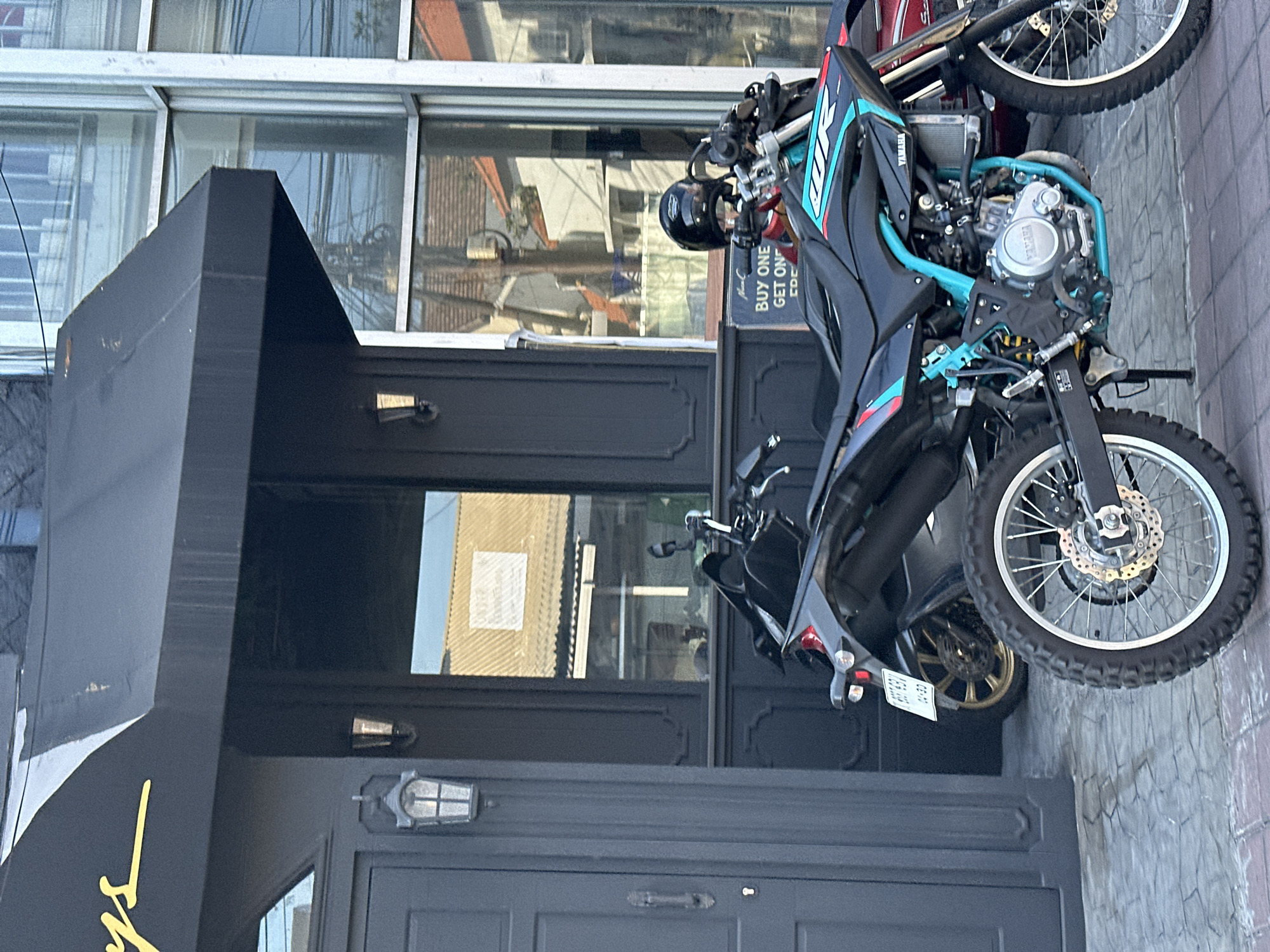 In North Kuta, Indonesia, two motorcycles are parked on a paved sidewalk in front of a building. The building features a black awning with yellow lettering that is partially visible at the top left. A dark gray door is to the left of the motorcycles. The motorcycle in the foreground is black with turquoise and red accents, and has "WR" and "YAMAHA" branding visible. Its front wheel is prominent. Behind it, another motorcycle is mostly obscured, with its rear wheel and license plate visible. A black helmet rests on the handlebars of the foreground motorcycle. A sign with "BUY ONE GET ONE" printed in white text on a blue background is also visible. The environment suggests a commercial area with buildings and reflective windows displaying the surroundings.