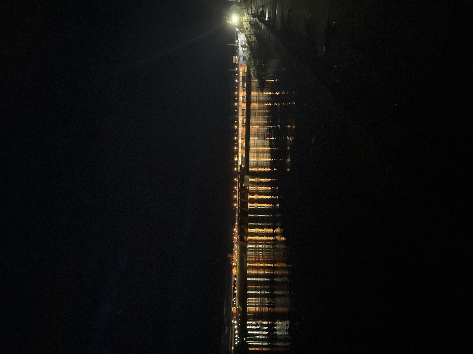 The image depicts a nighttime panoramic view of the Votkinsk Hydroelectric Power Station on the Kama River in Chaikovsky, Perm Krai, Russia. The scene is dominated by the power station's main structure, which stretches horizontally across the midground. The station is extensively illuminated by numerous warm-toned, likely orange or yellow, lights positioned along its length, creating a striking visual against the dark sky and water. These lights are reflected in elongated, shimmering streaks on the river's surface in the foreground, indicating subtle movement or ripples in the water.
The central part of the power station features a series of arches or gates, consistent with a dam or spillway, brightly lit from below. To the left of the main structure, a darker, elevated, and more complex industrial section of the power plant is visible, also adorned with scattered lights. The sky above is uniformly dark, suggesting a clear, moonless night or heavy cloud cover obscuring celestial bodies.
On the far right, a section of the riverbank or shoreline is partially visible, appearing as a dark, irregular strip. A singular, intensely bright white light source illuminates a small area on this bank, casting a glow that contrasts with the warmer lights of the power station. Indiscernible shadowy shapes or structures are faintly discernible near this bright light on the bank. The foreground consists entirely of the dark, reflective river water. No identifiable individuals or specific actions are discernible within the frame.