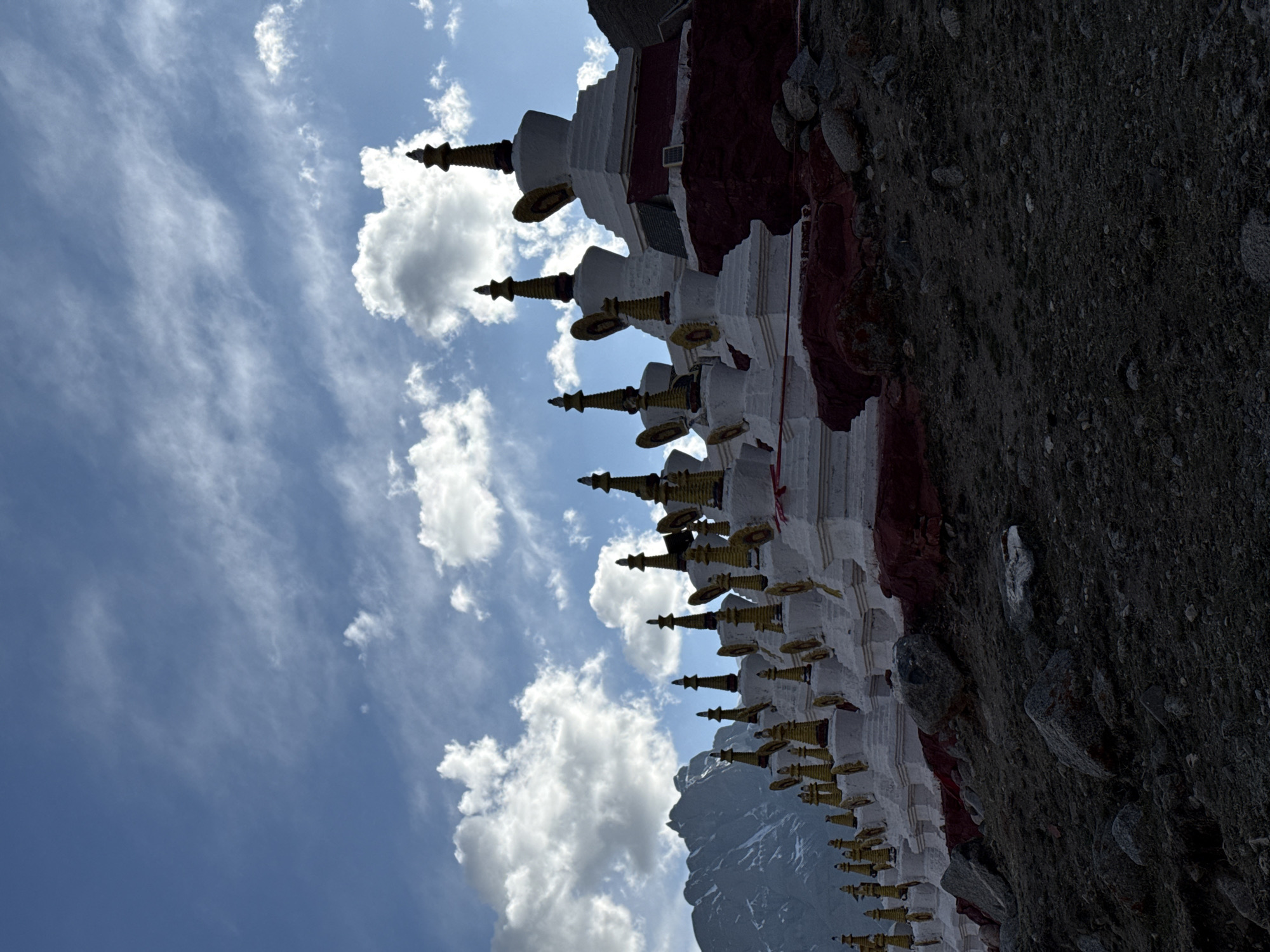 The image shows a row of white and red Buddhist stupas with golden spires aligned against a rocky terrain under a partly cloudy, blue sky. Snow-capped mountains are visible in the background. It is not possible to confirm the location within Parga town, China, based on the provided visual information.