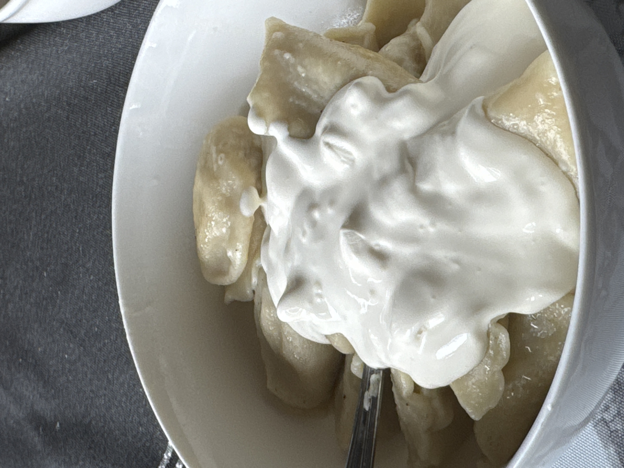 A close-up shot shows a white bowl filled with pierogi, a type of dumpling, generously topped with a dollop of sour cream. The pierogi are light yellow and have a slightly wrinkled texture. A portion of a silver fork is visible on the left side of the bowl, submerged among the pierogi. The bowl rests on a gray fabric surface, possibly a tablecloth or placemat. The background is a dark gray fabric with a textured pattern. The image appears to be taken indoors, likely in a home setting in Zaokskiy, Russia.