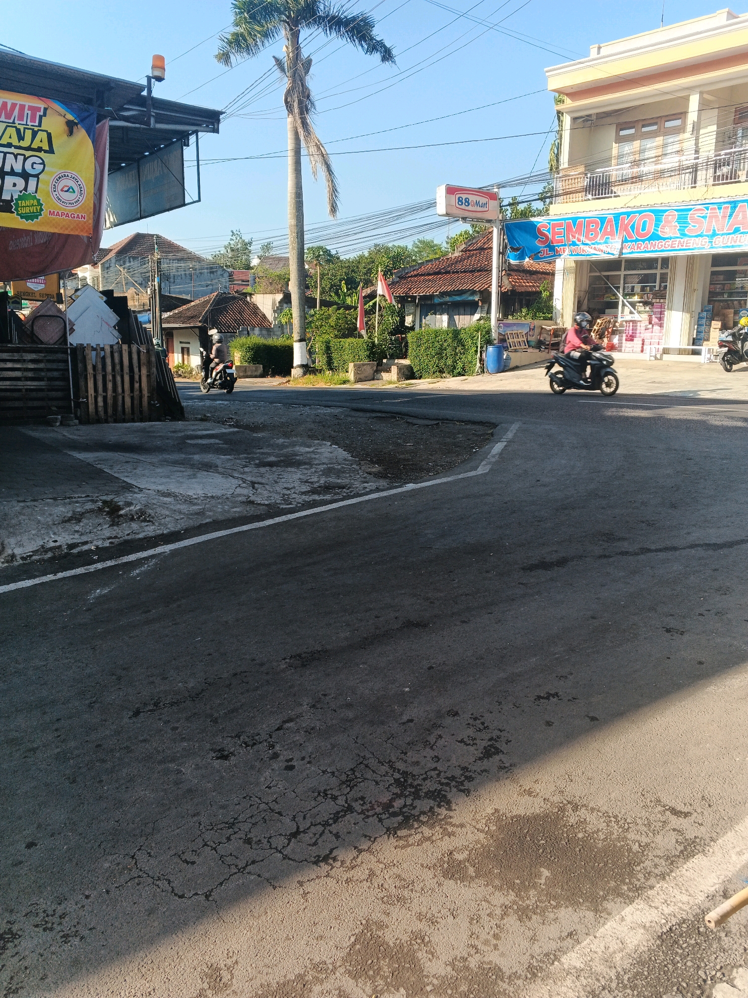 The image captures a street scene in Semarang, Indonesia, under a clear blue sky. In the foreground, a paved road is visible, showing signs of wear with dark patches and cracks. Sunlight casts a shadow across the road from the left.
On the left side of the image, part of a building with a dark roof is visible, adorned with a large yellow banner displaying text in Indonesian, including "WIT RAJA" and "NG SRI". Below the banner, there are stacks of wooden pallets and some metallic objects. A traffic light with an orange lamp is mounted on the roof.
Further down the street, a row of houses with red tile roofs is seen. A tall palm tree stands to the right of these houses, reaching towards the sky.
On the right side of the road, a two-story building houses "88 Mart" and a store named "SEMBKO & SNA" with a blue banner. A person is riding a black motorcycle, heading away from the camera, in front of the "SEMBKO & SNA" store. Another motorcycle is parked to the right of the store. Several blue plastic barrels are also visible near the store.
The overall impression is a typical street view in a moderately urban area of Semarang, Indonesia, during daylight hours.