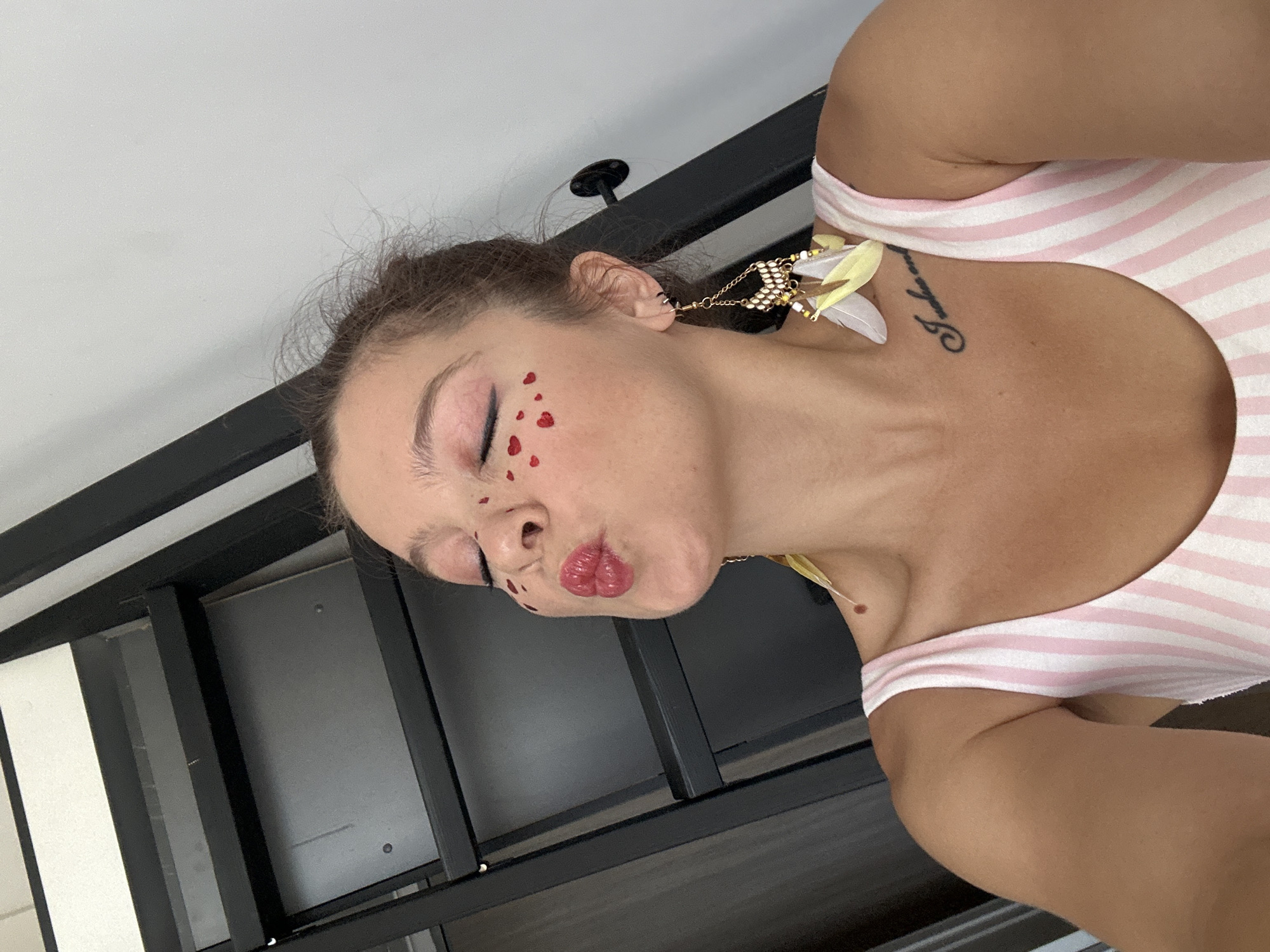 A woman is captured in a close-up, high-angle selfie taken indoors. She is leaning forward, puckering her lips as if to give a kiss, with her eyes closed. Her face is adorned with small red heart stickers scattered across her cheeks and forehead. She wears a pink and white striped swimsuit or top and is accessorized with a distinctive earring featuring gold chains, beads, and feathers. A tattoo is visible on her upper chest. The background is dominated by a black metal shelf unit, suggesting a home or studio environment. The lighting is bright, illuminating the subject and her features. The overall impression is one of playful self-expression.