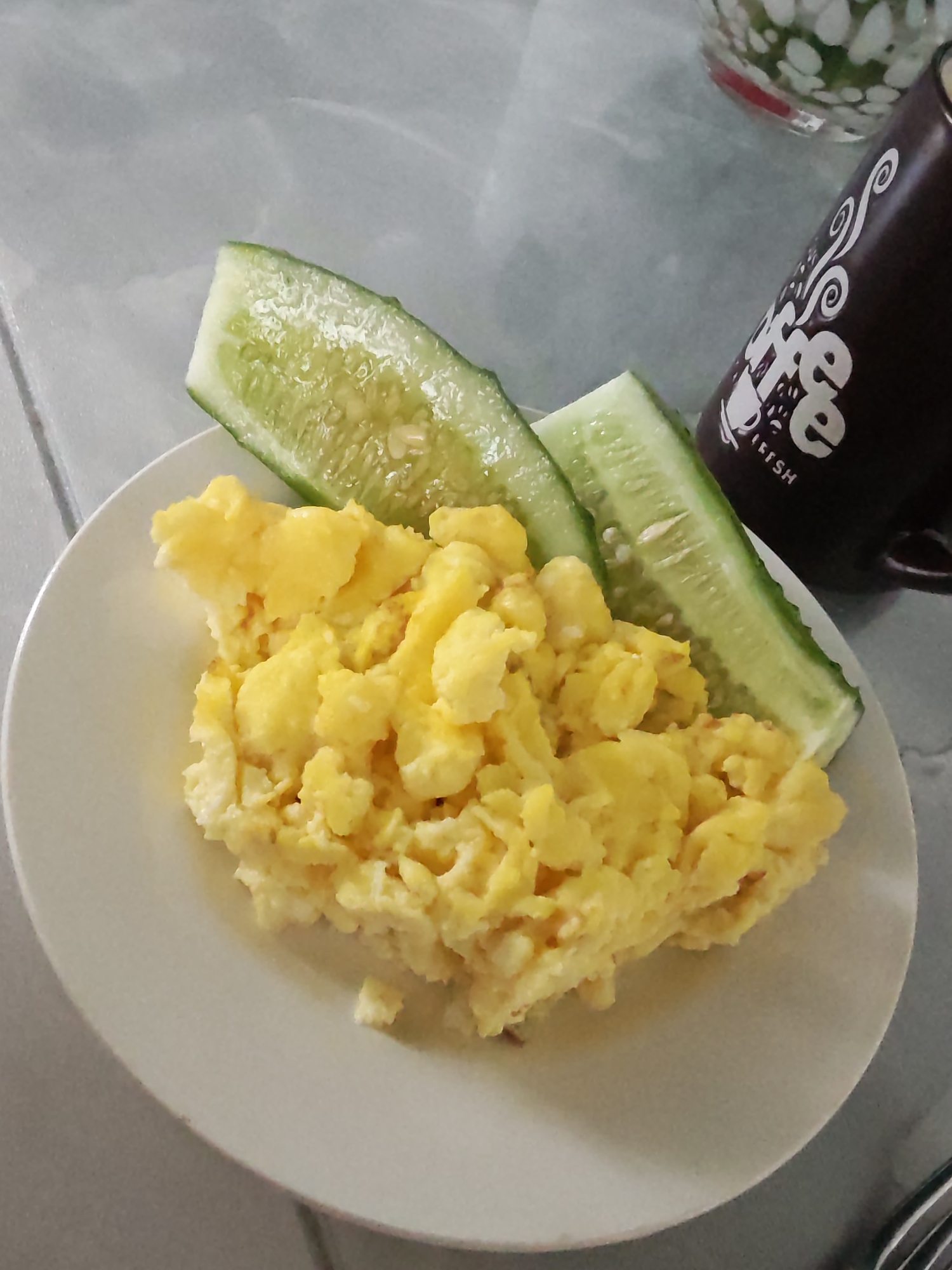 A top-down shot shows a white plate with scrambled eggs and two sliced cucumbers next to a dark brown mug with a white design. A fork is partially visible in the lower right. The background is a textured light gray surface. The image was likely taken indoors. There is no information that locates it in the city of Rybolovo, Russia. There is no information about people, actions, interactions, or the environment besides what is visible.