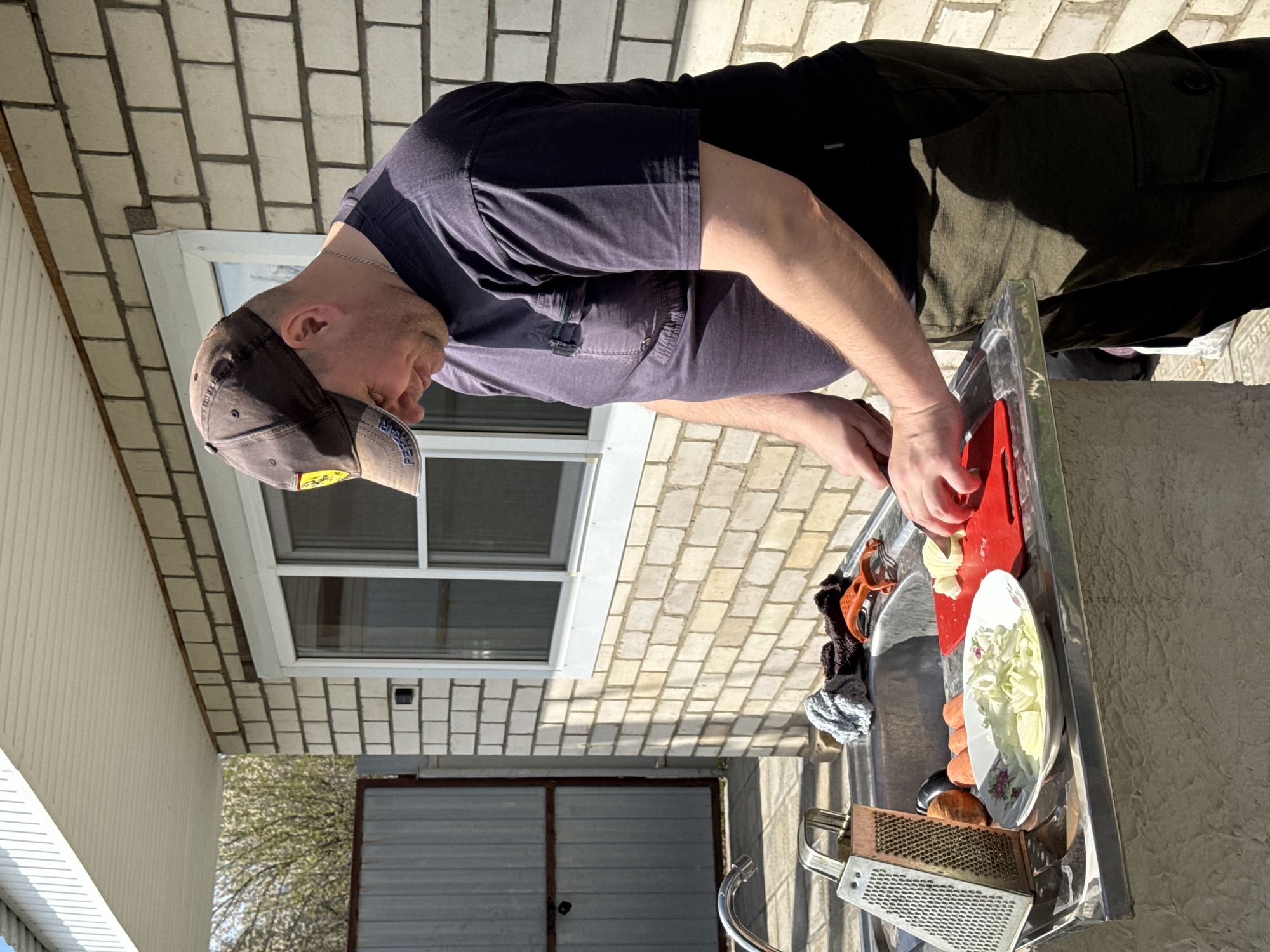 A man wearing a dark cap and T-shirt is cutting food on a red cutting board outdoors. The cutting board rests on a metal surface next to a white plate filled with leafy greens and a metal grater. The man is outside near a brick wall with a white-framed window. A metal garage door is visible in the background.