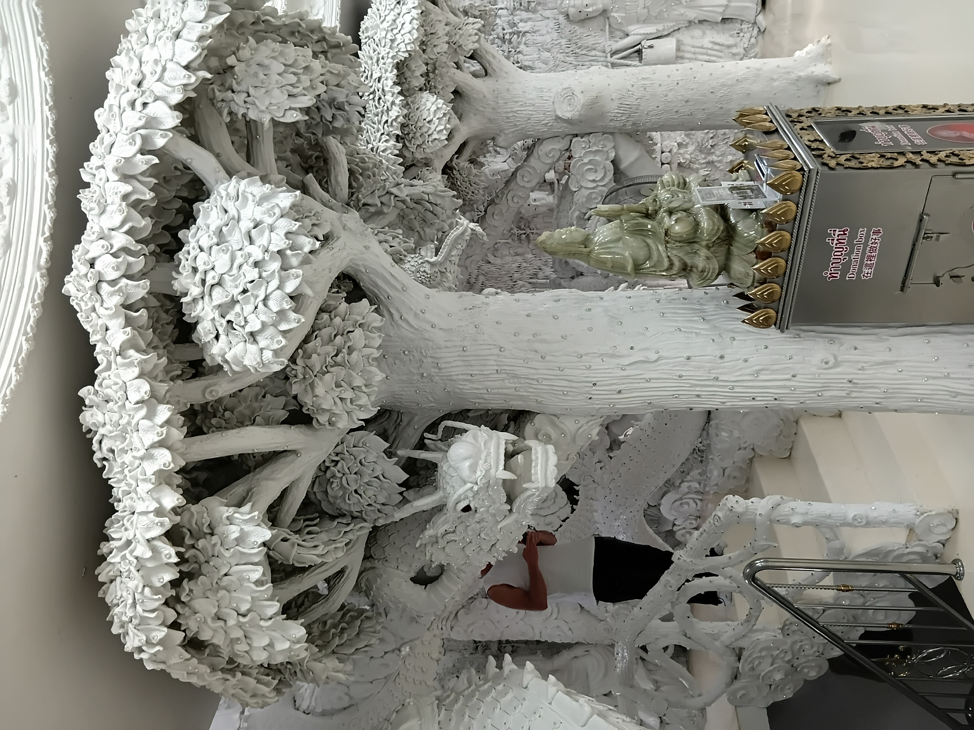 A person wearing a white sleeveless shirt and dark pants stands partially obscured behind a large, white, intricately sculpted dragon. The dragon is positioned on the left side of the frame, with its head and neck visible, featuring sharp teeth and horns. Behind and around the dragon are white sculptures of trees with dense foliage and numerous small, sparkling embellishments. To the right of the dragon, a staircase with a silver railing leads upwards. In the foreground and center, there is a donation box with gold decorative elements on top and ornate frames. A pale green, seated statue of a figure is placed behind the donation box. The entire scene is rendered in white, creating a monochromatic and detailed artistic environment, likely a temple or art installation in Ban Lao Phatthana, Thailand.