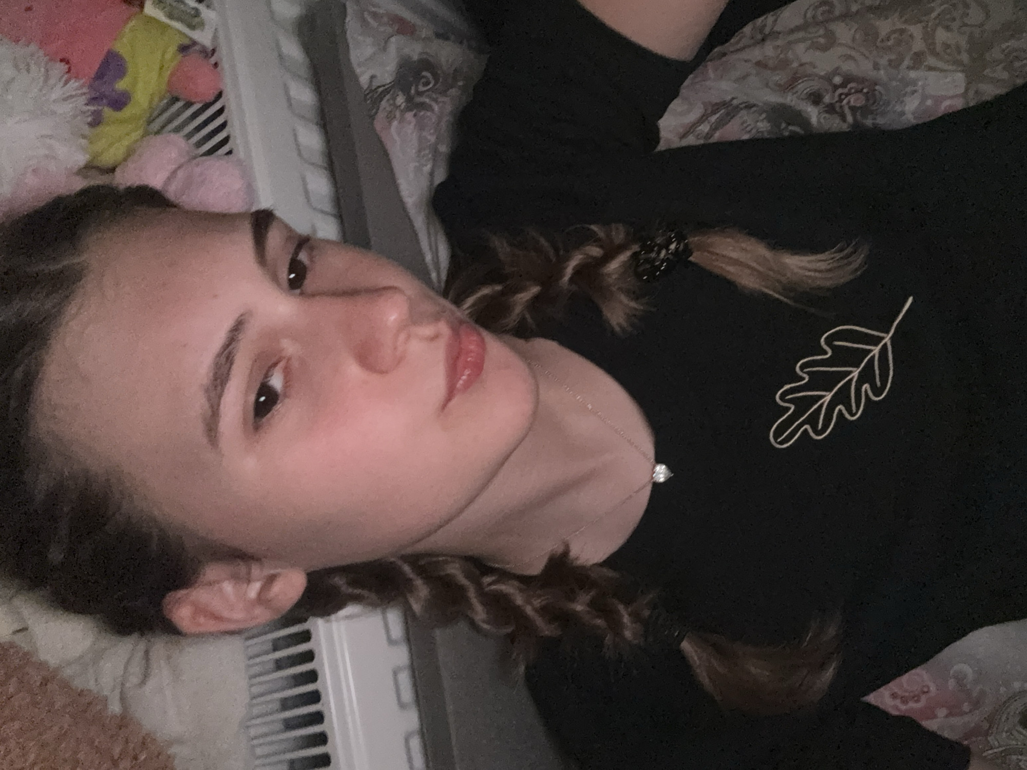 An indoor portrait features a young person, likely female, lying down and looking directly towards the camera. The image is taken from a slightly elevated perspective, with the subject positioned horizontally across the frame. She has medium-brown hair styled in two braids, one visible over her left shoulder (viewer's right) and secured with a black, sparkly hair tie. Her expression is neutral, with dark eyes and slightly parted lips. She is wearing a black long-sleeved top adorned with a distinctive outline of an oak leaf design in a lighter color on her left chest. A delicate silver-colored necklace with a small, sparkling pendant is visible around her neck.
The background suggests a cozy, indoor setting, likely a bedroom or living room in Panagyurishte, Bulgaria. To the upper left of the frame, a white radiator with vertical slats is partially visible. Above the subject's head, various soft objects can be seen, including a white fluffy item, a yellow object with a purple motif, and two pink, spherical items, possibly stuffed toys or decorative pillows. The bedding or upholstery beneath and around her features a light-colored, intricate patterned design. The lighting appears soft and indirect, typical of indoor artificial light, contributing to a calm and casual atmosphere. No discernible text is visible in the image.
