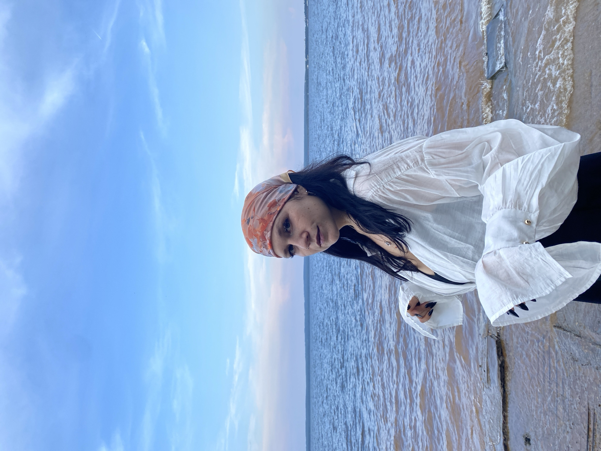 A young woman with dark hair and a bandana with an orange and red pattern is standing on the shore of a body of water. She is wearing a white, flowing shirt with puffy sleeves and dark clothing underneath. Her hands are visible, with black nail polish. The water is a murky brown color and there are some concrete slabs or rocks near the shore. The sky is blue with scattered clouds, and a distant horizon is visible across the water. The location is the Kama River in Chaikovsky, Russia.