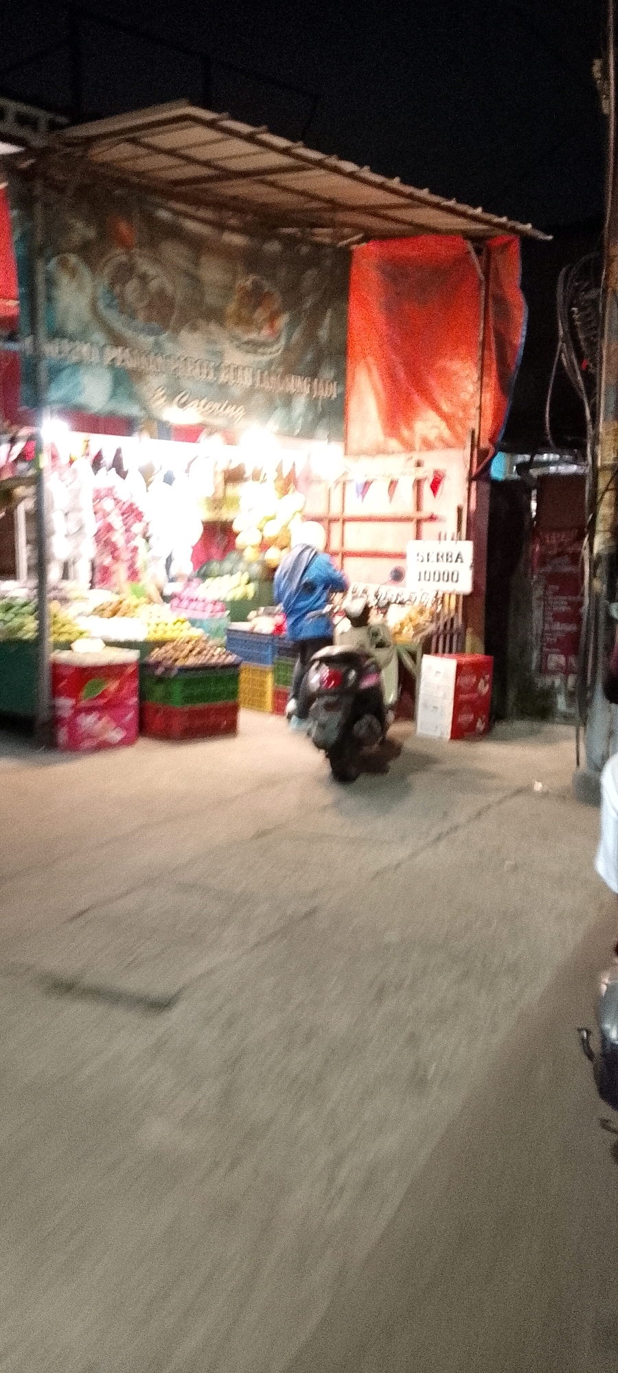 The image depicts a street scene at night in the city of Karangsatria, Indonesia, likely a marketplace or roadside stall area. A person wearing a blue jacket and a white head covering is visible standing next to a scooter, which is parked on the paved ground. The scooter is facing away from the viewer.
In the background, a stall is set up with a canopy, displaying various items that appear to be fruits and vegetables. Bright lights illuminate the stall, highlighting the produce. There are several crates filled with goods, in different colors including red, green, and blue. A banner with text and images hangs above the stall. To the right of the stall, a white sign with black text reading "SERBA 10000" is displayed, indicating a price point. Further to the right, a red and white box is visible. The overall environment is dimly lit, with the artificial lights from the stall being the primary light source. The ground is a rough, textured pavement.