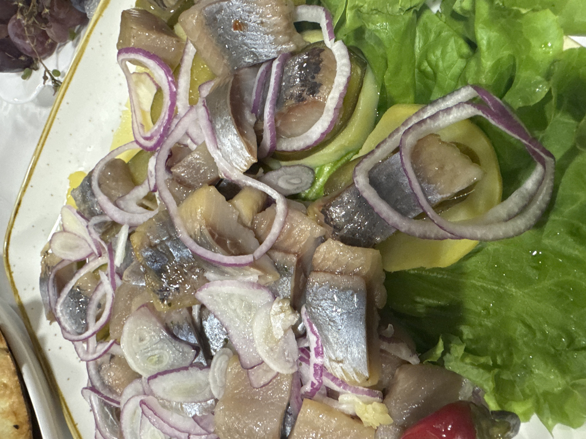 A close-up, overhead shot shows a serving of marinated herring with thinly sliced red onions and pickles, laid out on a bed of lettuce. The fish is cut into bite-sized pieces, with their silvery skin still attached. The onions are in rings, and slices of pickle are visible amongst the fish. The dish is served on a white plate with a thin yellow border, positioned on a table. In the upper left, a piece of bread or pastry is partially visible. In the background, on a separate plate, a bunch of dark grapes can be seen. The lighting is bright, indicating an indoor setting, possibly a restaurant. There is no indication of the specific location within Tash-Moinok, Kyrgyzstan.
