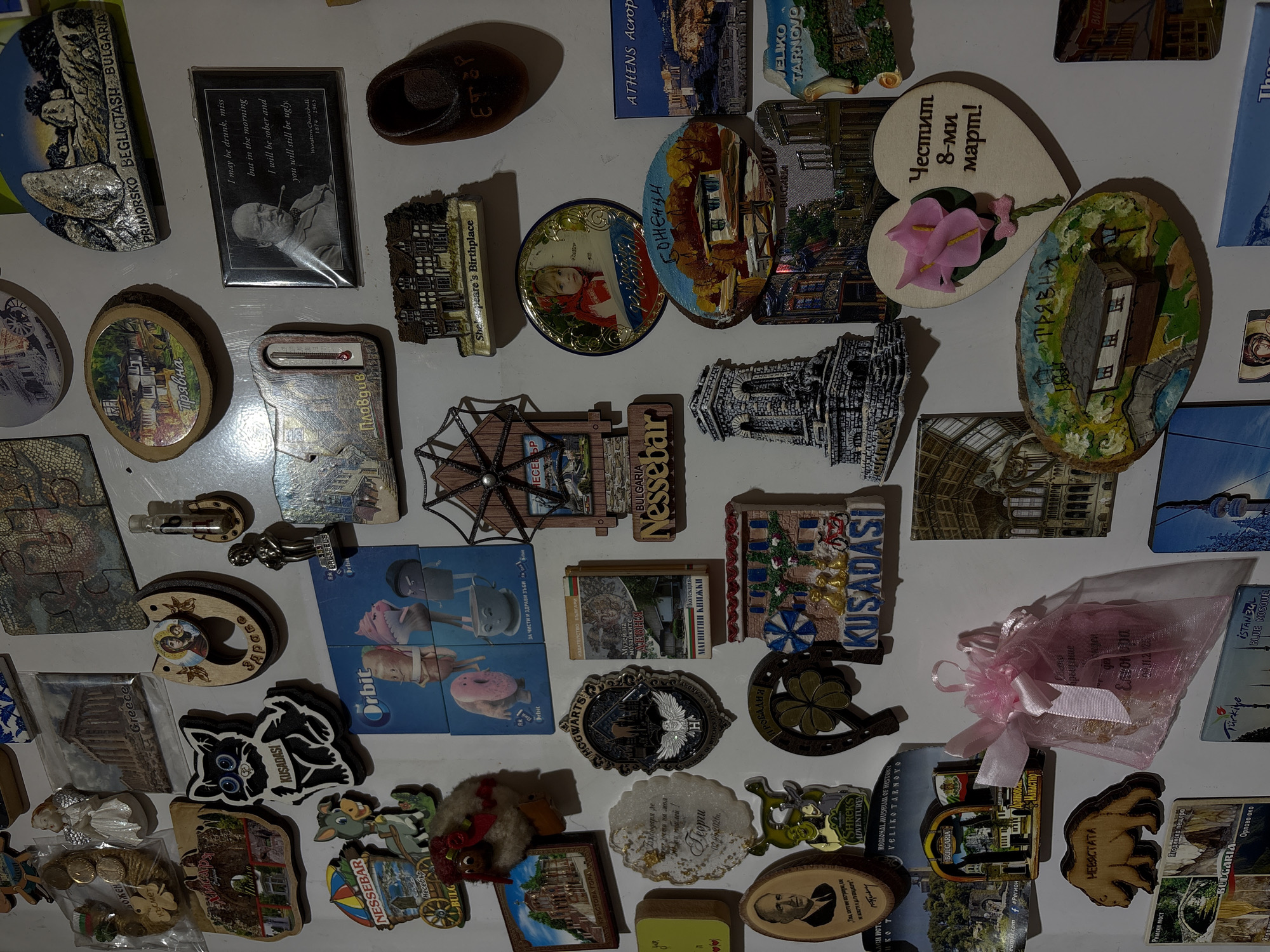 The image displays a collection of magnets and souvenirs affixed to a white wall, creating a cluttered yet vibrant arrangement. The items are diverse, featuring miniature buildings, scenic landscapes, cartoon characters, and quotes. Several magnets appear to represent Bulgarian locations, indicated by text such as "Nessebar," "Primorsko Beglitsash Bulgaria," and "Veliko Tarnovo." One magnet specifically mentions "Athens Acropolis." There is a prominent magnet with a portrait of Winston Churchill and a quote attributed to him. Another item is a wooden heart-shaped ornament with flowers and Bulgarian text that reads "Честит 8-ми март!" (Happy 8th of March!). Also visible is a wooden cutout of a bear with the text "НЕВЯСТАТА" (The Bride) below it. A small, pink, sheer bag tied with a pink ribbon contains what appear to be small items or trinkets. The overall impression is a personal collection of mementos from travels and gifts. There is no specific visual element that definitively places this collection within the city of Provadia, Bulgaria.