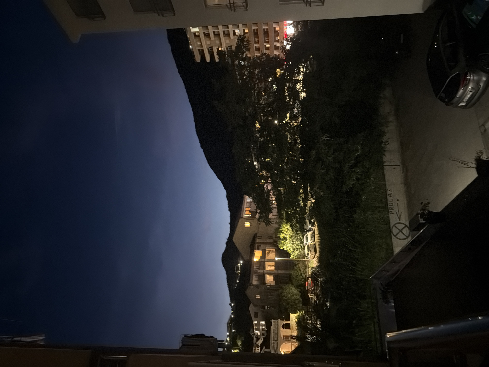 The image captures a nighttime view of a residential area in Budva, Montenegro, characterized by buildings nestled on a hillside against a dark, twilight sky. On the right, a modern apartment building with numerous lit windows stands prominently. To the left, several smaller buildings, some with illuminated windows, are spread across the slope. A dense cluster of trees with visible lights shining through them occupies the midground. In the foreground, a paved area appears to be a parking lot or driveway, with a silver hatchback parked on the right. A street sign indicating "PROLAZ" (meaning "passage" in Montenegrin) with an arrow and a circular prohibition sign is visible on the left side of the paved area. The overall atmosphere is tranquil, with the artificial lights of the buildings and streetlamps providing illumination in the darkness.