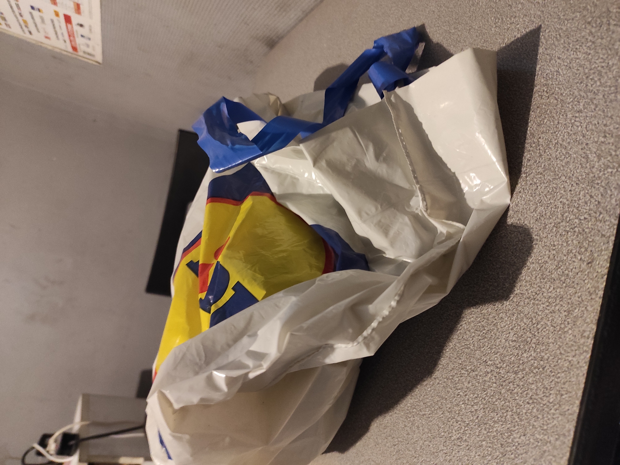 A crumpled white plastic shopping bag with blue and yellow branding, possibly from Lidl, lies on a speckled grey countertop. The bag appears to be empty and is partially collapsed, with its handles made of twisted blue plastic. The background consists of a grey wall and a dark surface that may be a chair or table. To the left, a power strip with cords is visible, plugged into an outlet on a white box. A poster with text and icons is affixed to the wall in the upper right corner. The scene suggests an indoor setting, likely a home or commercial establishment, with no people present. The lighting is dim, casting shadows on the countertop. The image was taken in Chaponost, France.