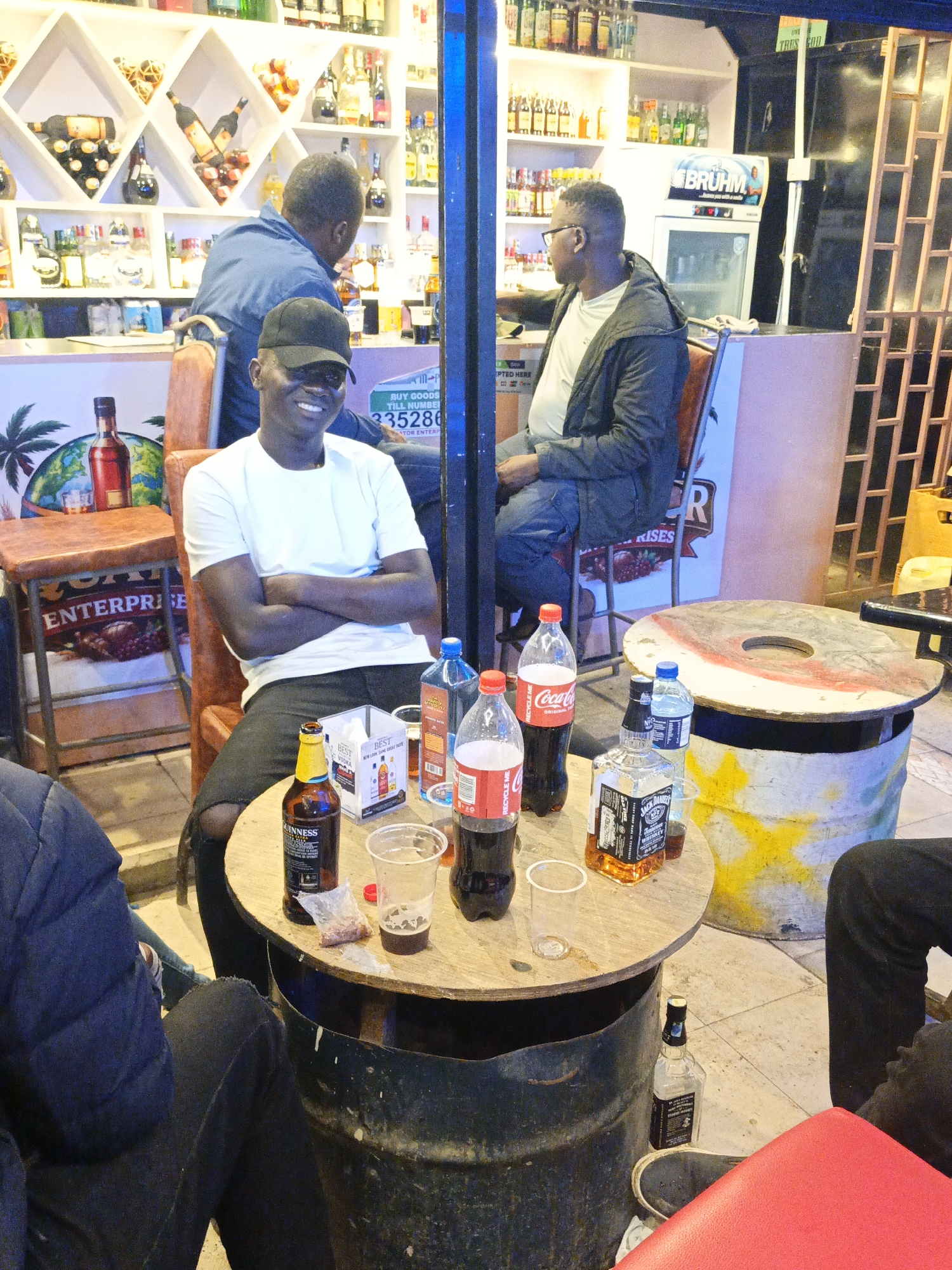 The image captures a casual social scene at what appears to be an outdoor bar or pub in Nairobi, Kenya. Several people are present. A smiling man in a white t-shirt and black cap sits at a table, arms crossed. Two other men are seated at the bar behind him, engaged in conversation. Another man is partially visible on the left, and a fourth man is partially visible to the right, also sitting at the table.
The table is laden with bottles and drinks, including bottles of Guinness, Coca-Cola, and Jack Daniel's whiskey, along with bottles of water and a small bag with meat snacks. The table itself is made from a repurposed metal drum. There is a second table nearby, similarly constructed.
Behind the table, the bar area is well-stocked with various alcoholic beverages displayed on shelves and in a cooler. A sign displays a local phone number, advertising goods.