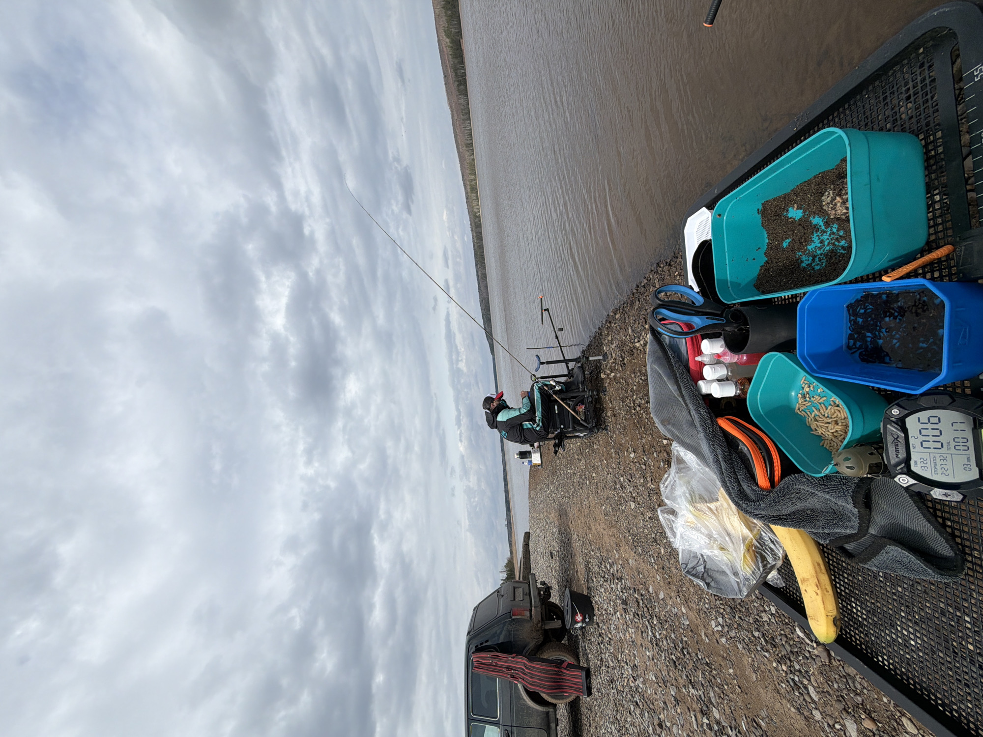 The image depicts a fishing scene, possibly in the city of Chaykovsky, Russia, given the presence of a Russian-made Lada Niva vehicle. A person is seated in a fishing chair near the water's edge, with a fishing rod set up. The water appears brown and murky, with a forested shoreline visible in the distance. The sky is overcast with grey clouds. A tray with various fishing supplies sits nearby, including containers of bait (maggots, groundbait, and worms), a banana, and a digital watch. The ground is covered with pebbles. A Lada Niva vehicle is parked close to the fishing spot.