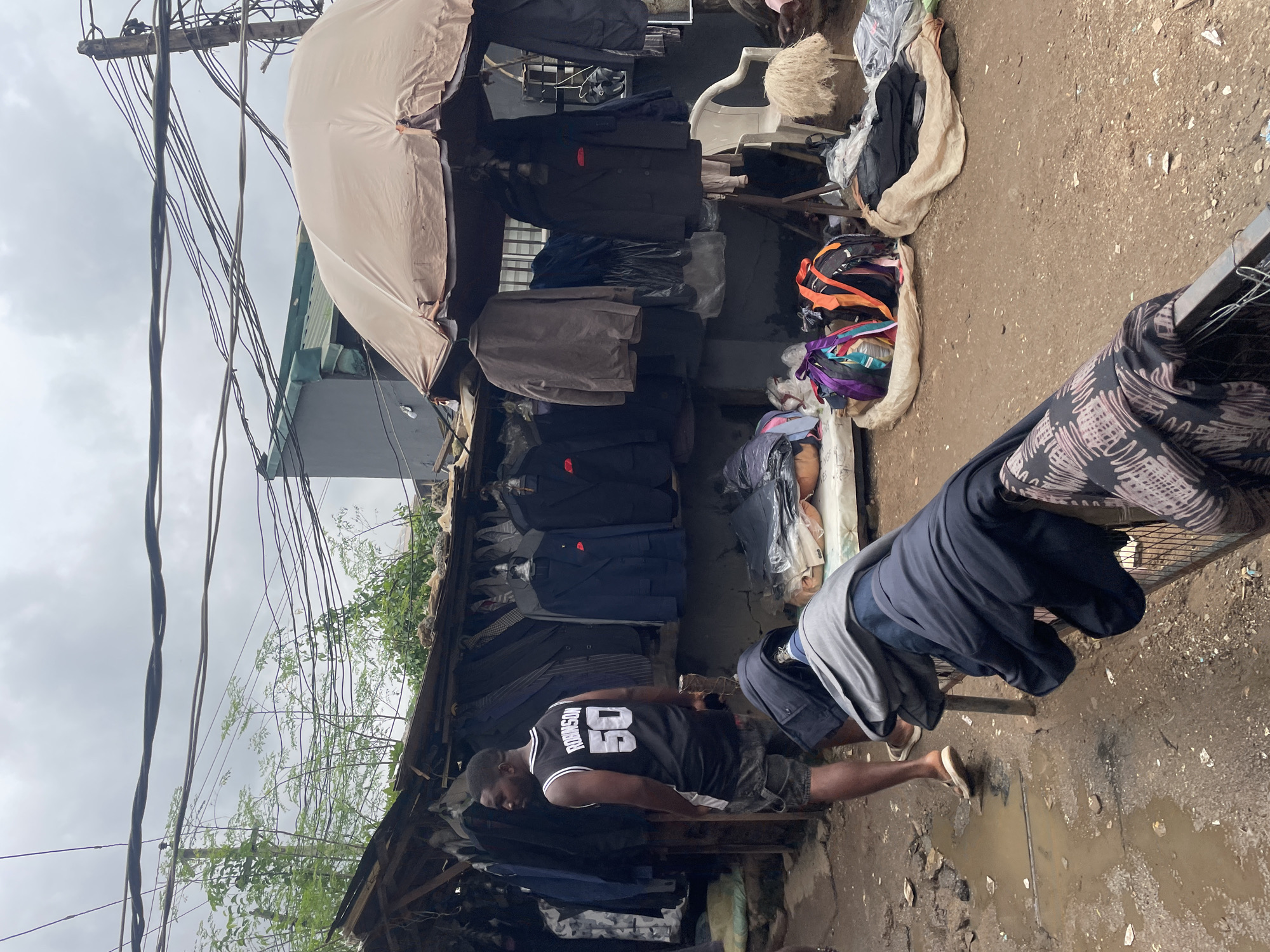 The image depicts an outdoor clothing market stall in Abuja, Nigeria, characterized by a somewhat disorganized setup. A man wearing a black basketball jersey with "ROBINSON 50" on the back is standing and looking to his left, dressed in denim shorts. In the foreground, various garments are draped over a metal railing, including a dark navy jacket and a black and pink patterned fabric. Behind the man, a stall displays a collection of men's suits and jackets hanging on a rack, with a large beige umbrella providing shade over some of the clothing. Several bags, some colorful and others black, are placed on the ground. The ground surface is a mix of dirt and puddles, suggesting recent rain or a damp environment. Overhead, a tangle of electrical wires runs across the frame, indicating a dense urban infrastructure. A white plastic chair is visible near the stall, and a pile of light-colored, shaggy material is on the ground to its right. The overall scene suggests a busy, informal retail environment.