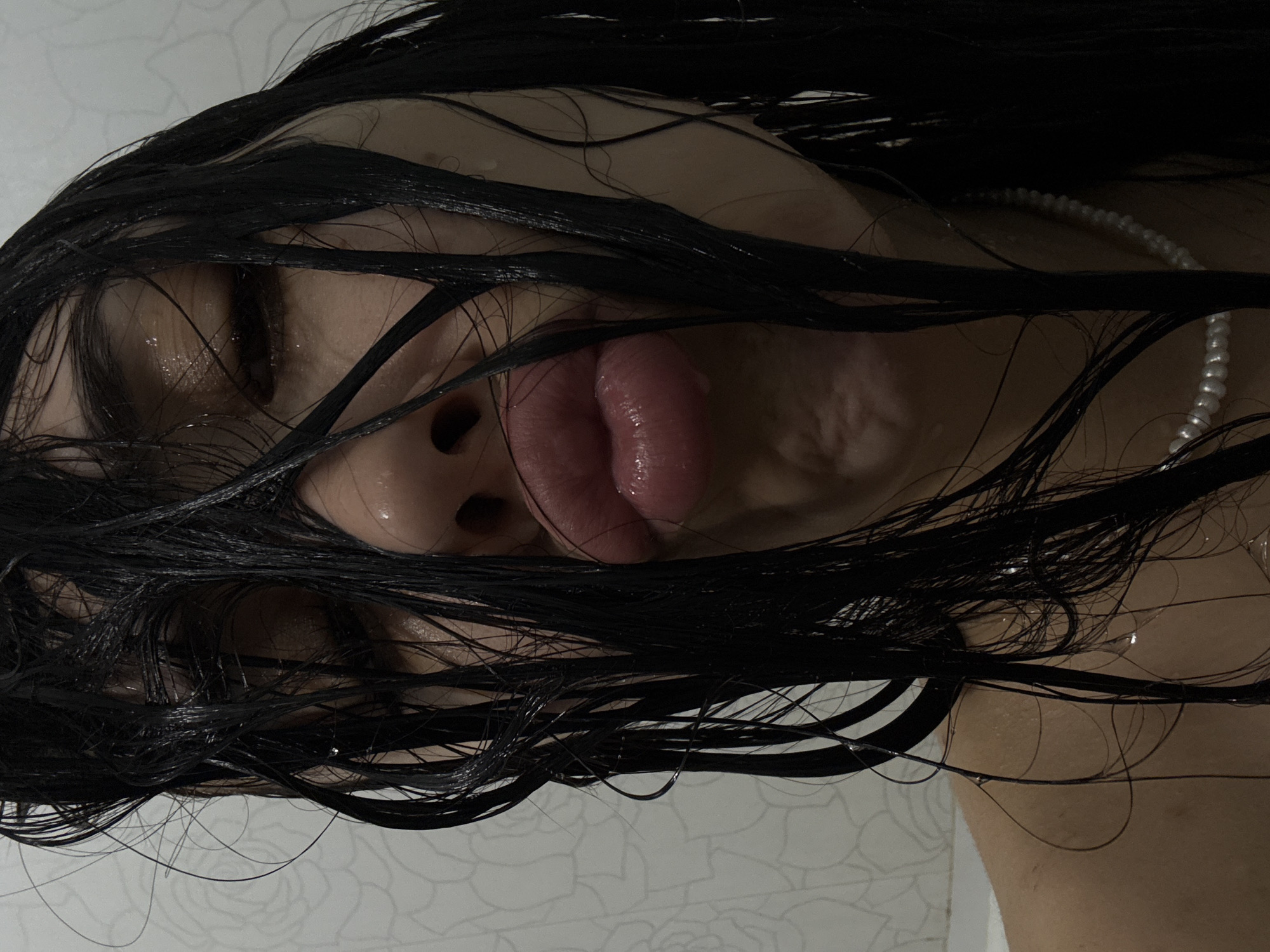 A close-up, inverted shot of a person with long, wet black hair covering their face and shoulders. Their eyes are closed, and their tongue is sticking out. A pearl necklace is visible on their neck. The background is a white tiled wall with a faint floral pattern. There is no information to suggest that this photograph was taken in Votkinsk, Russia.