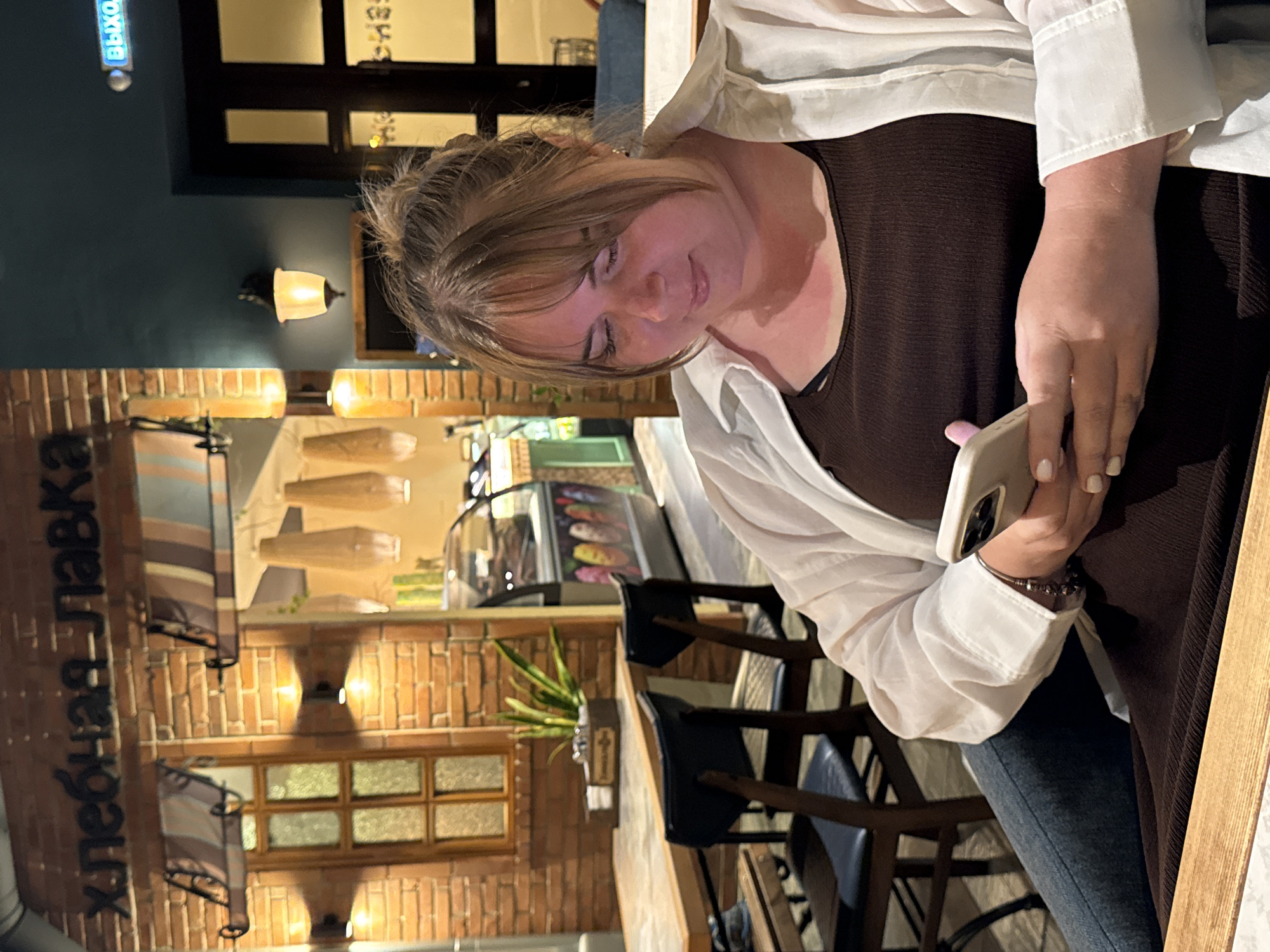 The image shows a woman sitting at a table in a dimly lit establishment, possibly a cafe or restaurant. She is wearing a dark brown top and an unbuttoned white shirt over it, paired with dark blue jeans. The woman is looking down at her phone, which she holds in her right hand. Her hair is styled loosely, with bangs framing her face, and she appears to be engrossed in her device.
The background suggests an indoor setting with a rustic theme. A brick wall is visible, adorned with several small, illuminated wall lamps. To the left, a large brown sign with white vertical text is partially visible, appearing to read "ХЛЕБНАЯ ЛАВКА" (Khlebnaya Lavka), which translates to "Bread Shop". Beyond the brick wall, there are reflections on glass, and what appears to be a display case with colorful items, possibly pastries or desserts. Wooden chairs and tables are also part of the scene.
The lighting is warm and intimate, suggesting it might be evening or nighttime. There are no indications of the weather outside. The overall atmosphere is relaxed and casual, with the woman appearing comfortable and absorbed in her phone. No other people are clearly visible in the frame, though the presence of chairs suggests it could be a public space. The image is oriented vertically, with the top of the woman's head towards the right side of the frame.