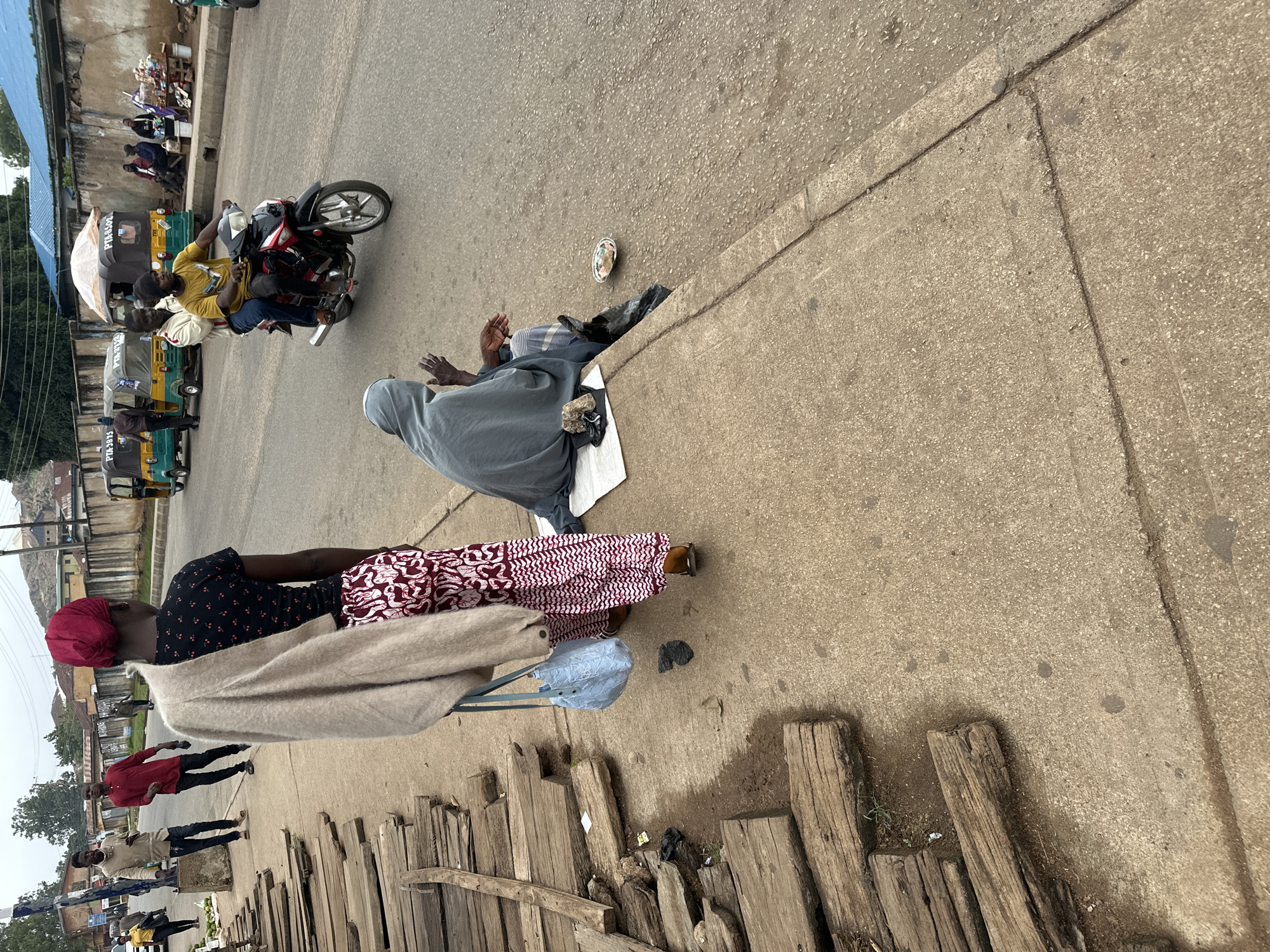 In the city of Unknown, Unknown, on a cloudy day, a street scene unfolds. A woman wearing a red headscarf and a patterned skirt stands facing away from the camera, a light-colored cardigan draped over her shoulders and a blue bag in her left hand. To her right, a person wrapped in a gray shawl sits on the ground, their hands held up in a gesture of supplication, with a small plate of items and a black plastic bag nearby. In the background, several people are visible. Two men walk on the sidewalk, one in a red shirt and the other in a light-colored jacket. Further down the street, a motorcycle carries two passengers. Several auto-rickshaws, known as 'Keke Napep' in Nigeria, are parked on the road, some with visible license plates. The street is paved with asphalt, and a concrete sidewalk runs along the edge, lined with stacks of weathered wooden planks. The environment appears to be a bustling urban area with buildings and vegetation visible in the distance.