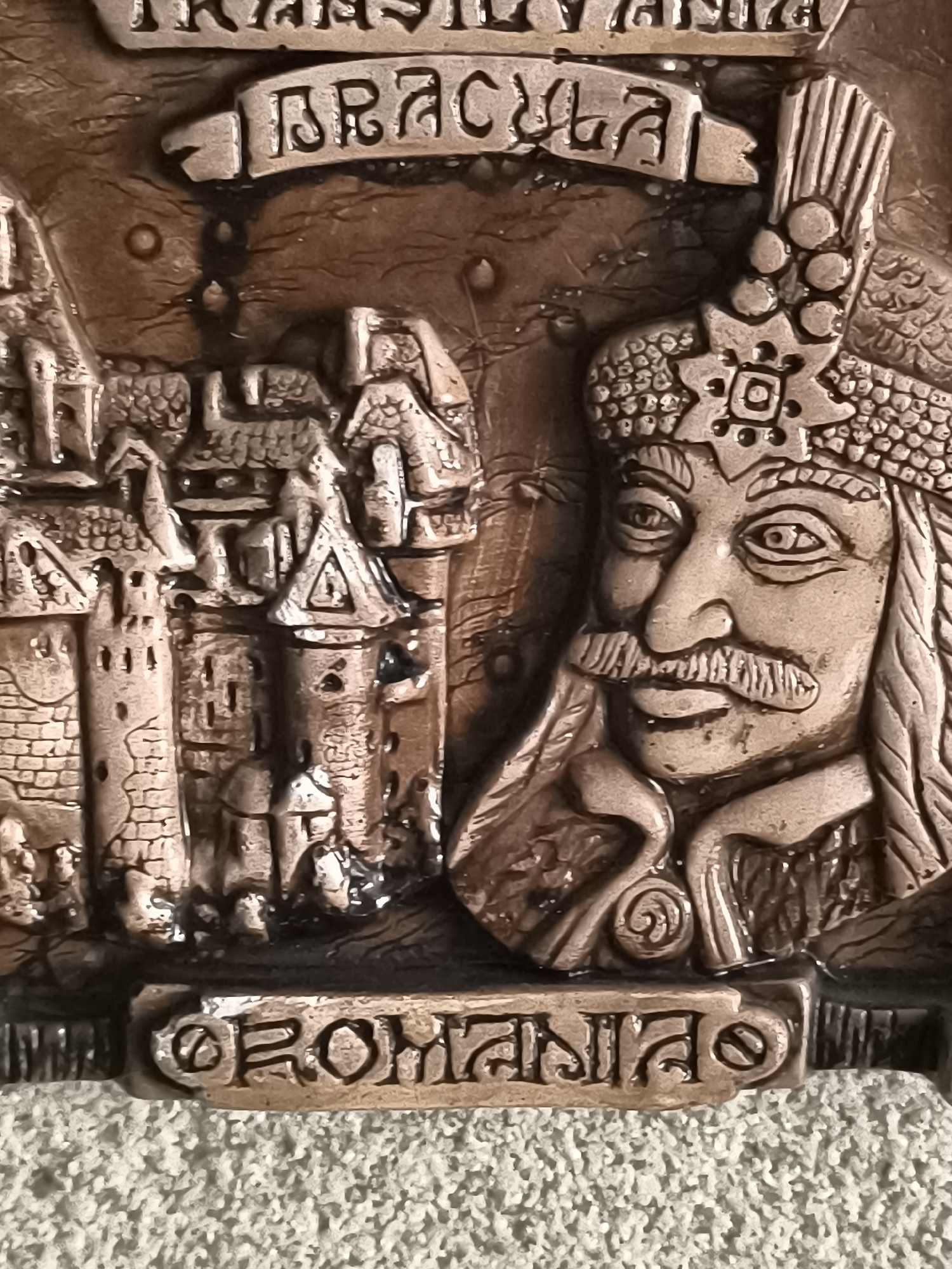 The image shows a close-up of a bronze-colored relief depicting a castle on the left and a man's face on the right. The man has a mustache and wears a headdress. Above the man's head, a banner reads "DRACULA". Above this, another banner is partially visible and appears to read "TRANSYLVANIA". Below the castle and the man's head, another banner reads "ROMANIA". The background is textured and appears to be a rough, uneven surface. The lighting highlights the raised details of the relief. The scene is not located in Dobrogea Veche, Moldova.