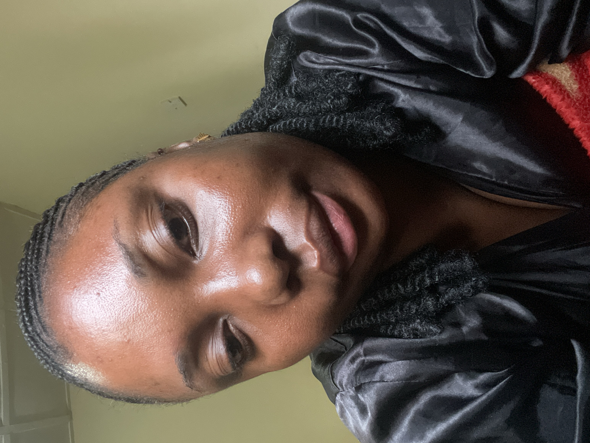 A close-up portrait of a young Black person, likely female, captured indoors in Jos, Nigeria. The image is rotated 90 degrees counter-clockwise, showing the subject's head tilted to the left, possibly indicating a selfie or a casual shot from a low angle. The person has dark skin with a natural, healthy sheen, particularly noticeable on the forehead, cheeks, and nose. Small, neat braids frame the hairline, and textured, curly black hair is visible peeking out from under a shiny black, satin-like garment, which appears to be a robe or sleepwear.
The subject has a relaxed expression, with a subtle, gentle smile playing on their lips. Their eyes are partially obscured by the angle, conveying a soft gaze. The background consists of a plain, light greenish-yellow wall or ceiling, providing a minimalist backdrop. On the far right, behind the subject's shoulder, a small section of a red and beige textured fabric, possibly a blanket or another item of clothing, is visible. The lighting appears soft and natural, highlighting the skin's contours and texture. There is no visible text in the image. The overall impression is one of calm and natural beauty within an indoor setting.