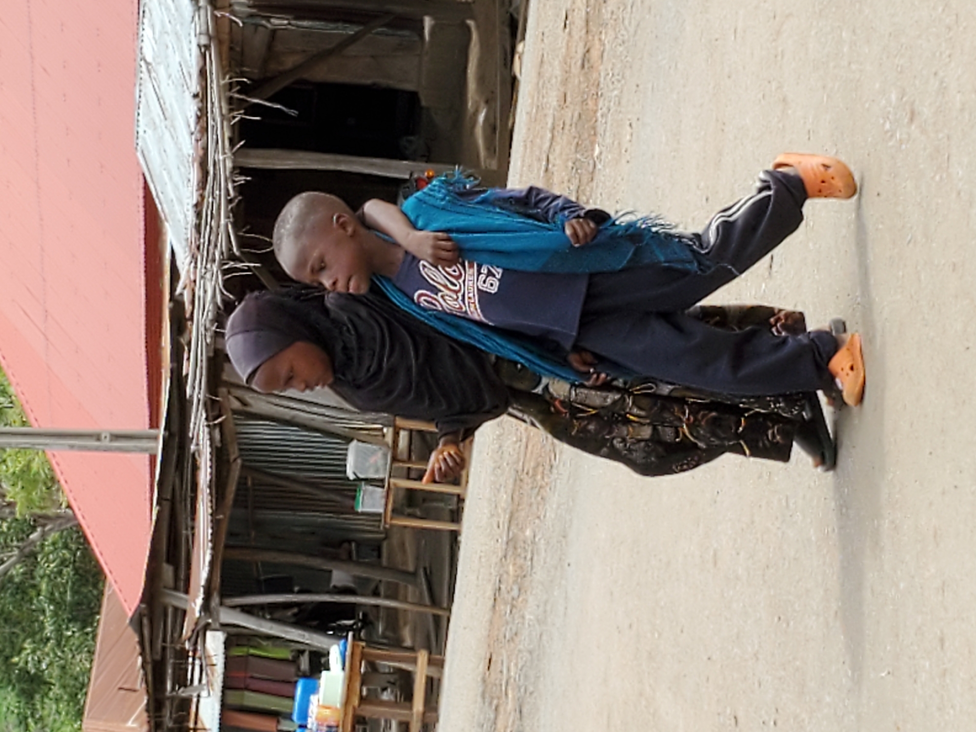 In Mararaba, Nigeria, a woman wearing a black head covering and a dark garment carries a young boy on her back. The boy is wearing a blue t-shirt, dark pants, and orange Crocs. A blue shawl is draped over him. They are standing on a concrete surface near a building with a rusty metal roof. Miscellaneous items, including containers and furniture, are visible behind them.