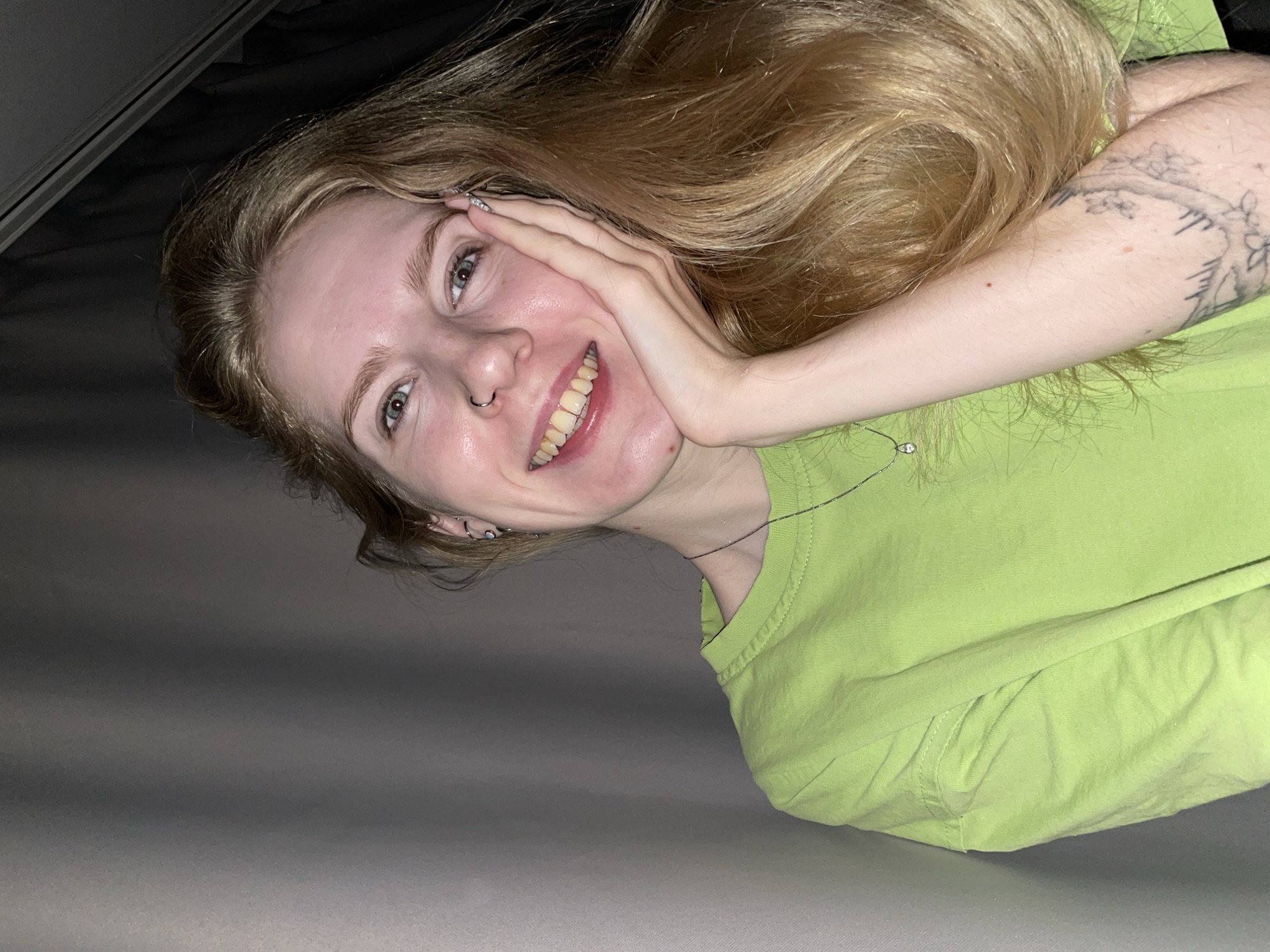 A young woman with long, wavy blonde hair lies on a gray surface. She is wearing a bright green t-shirt. She has fair skin, light green eyes, and a nose ring. Her left hand is touching her face as she smiles. There is a tattoo visible on her right arm. The upper left corner of the image is dark, suggesting she might be under something. The location is not discernible from the image.