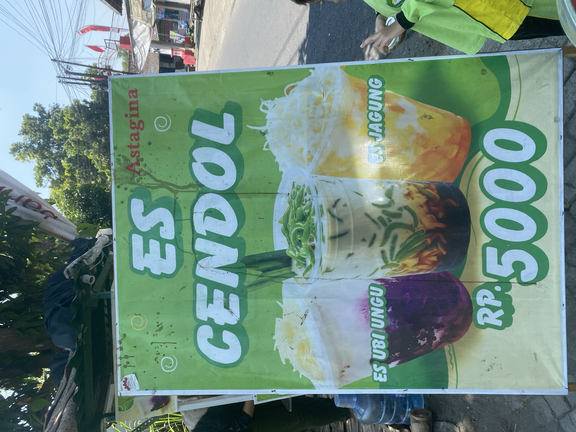 The image shows a large green banner advertising "ES CENDOL Astagina" with pictures of three drinks. The banner is placed outdoors, possibly on a street or sidewalk, with a road visible in the background. In the foreground, a person wearing a green and yellow outfit is partially visible on the right, and a large blue water dispenser is on the left. The banner itself is the main subject, with text and images related to the drinks. The environment suggests a commercial or food stall setting.