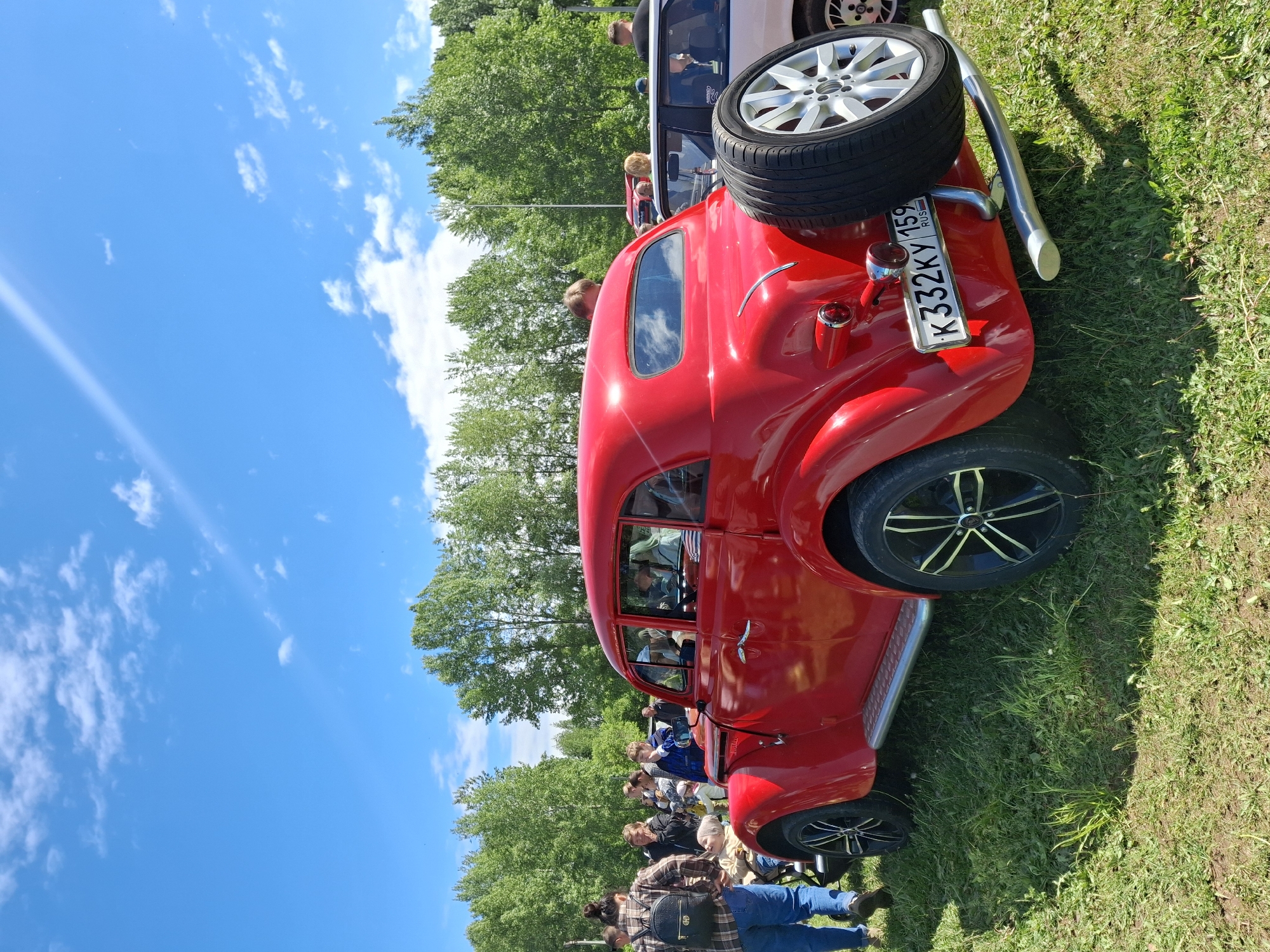 The image captures an outdoor scene on a sunny day, likely in Chaikovsky, Russia, as indicated by the license plate "K332KY 159 RUS". The primary subject is a bright red vintage or customized car, with a spare tire mounted on the back. The car is parked on a grassy field. A group of people, including men and women of varying ages, is present in the background, appearing to be looking at or admiring the car. They are casually dressed. The sky is clear blue with scattered white clouds. Trees are visible in the background. The ground is covered with green grass.