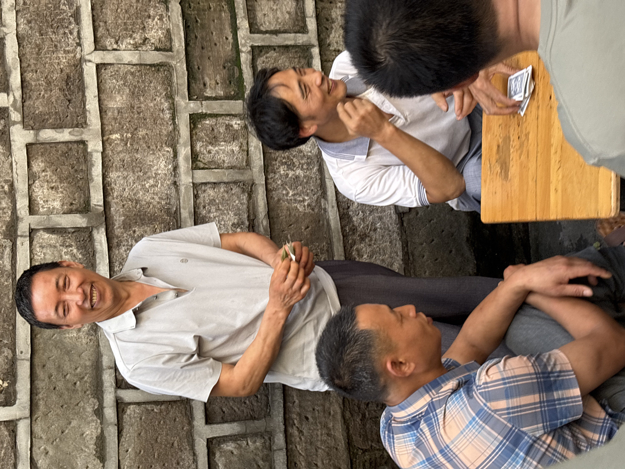 In Yuzhong District, China, four men are gathered around a wooden table, engaged in a game of cards. The man standing in the background, wearing a light grey polo shirt, is smiling and holding several cards in his hand. To his right, a man in a white polo shirt with grey trim is also smiling and appears to be holding cards. In the foreground, two men are seated. The man on the left wears a blue and pink plaid shirt and grey pants, his arms resting on his knees. The man on the right has his back to the camera, wearing a grey t-shirt. A deck of cards is visible on the wooden table between the seated men. The scene is set against a rough, textured stone wall.