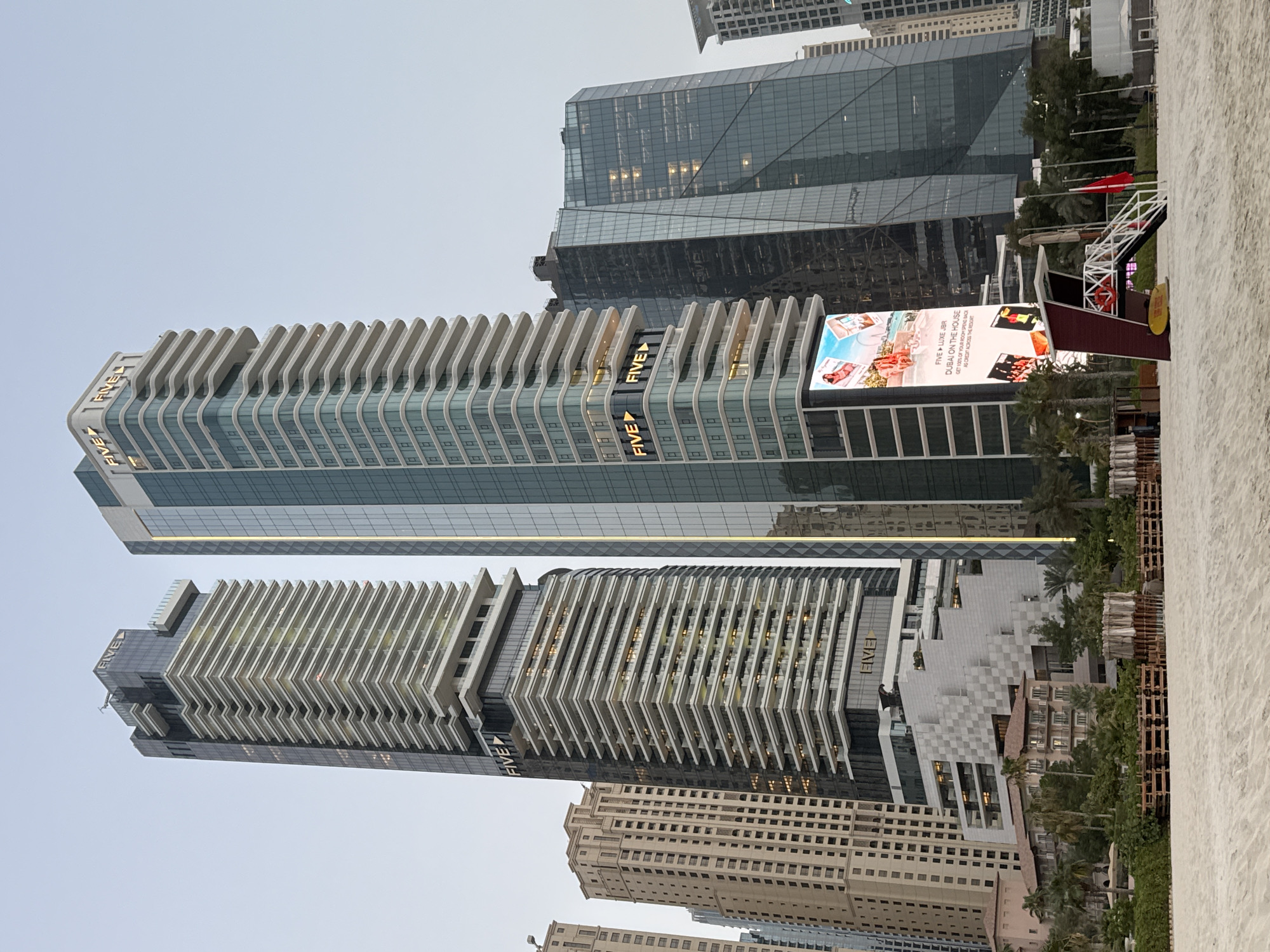 The image captures a view of the Jumeirah Beach Residence (JBR) area in Dubai, United Arab Emirates, with a focus on the FIVE Jumeirah Village hotel towers. In the foreground, a sandy beach is visible, sparsely populated with beach chairs and umbrellas. A red and white lifeguard station with a yellow surfboard rests on the sand to the right. Behind the beach, lush green vegetation and palm trees line the area, obscuring much of the lower sections of the buildings.
The dominant features of the image are two prominent skyscrapers. The left tower is a tall, slender building with a dark glass facade on its sides and numerous horizontal white balconies stacked one above another, creating a layered effect. The word "FIVE" is illuminated in gold lettering on the upper section of this building. The right tower is also tall and features a modern design with a primarily glass exterior and curving white balconies. The word "FIVE" is also illuminated in gold on this building, with two instances visible. In front of the right tower, a large digital billboard displays an advertisement featuring people on a beach. Other high-rise buildings, characterized by their glass facades and varying architectural styles, are visible in the background, creating a dense urban skyline. The sky is overcast, with a pale, diffused light, suggesting either early morning or late afternoon.