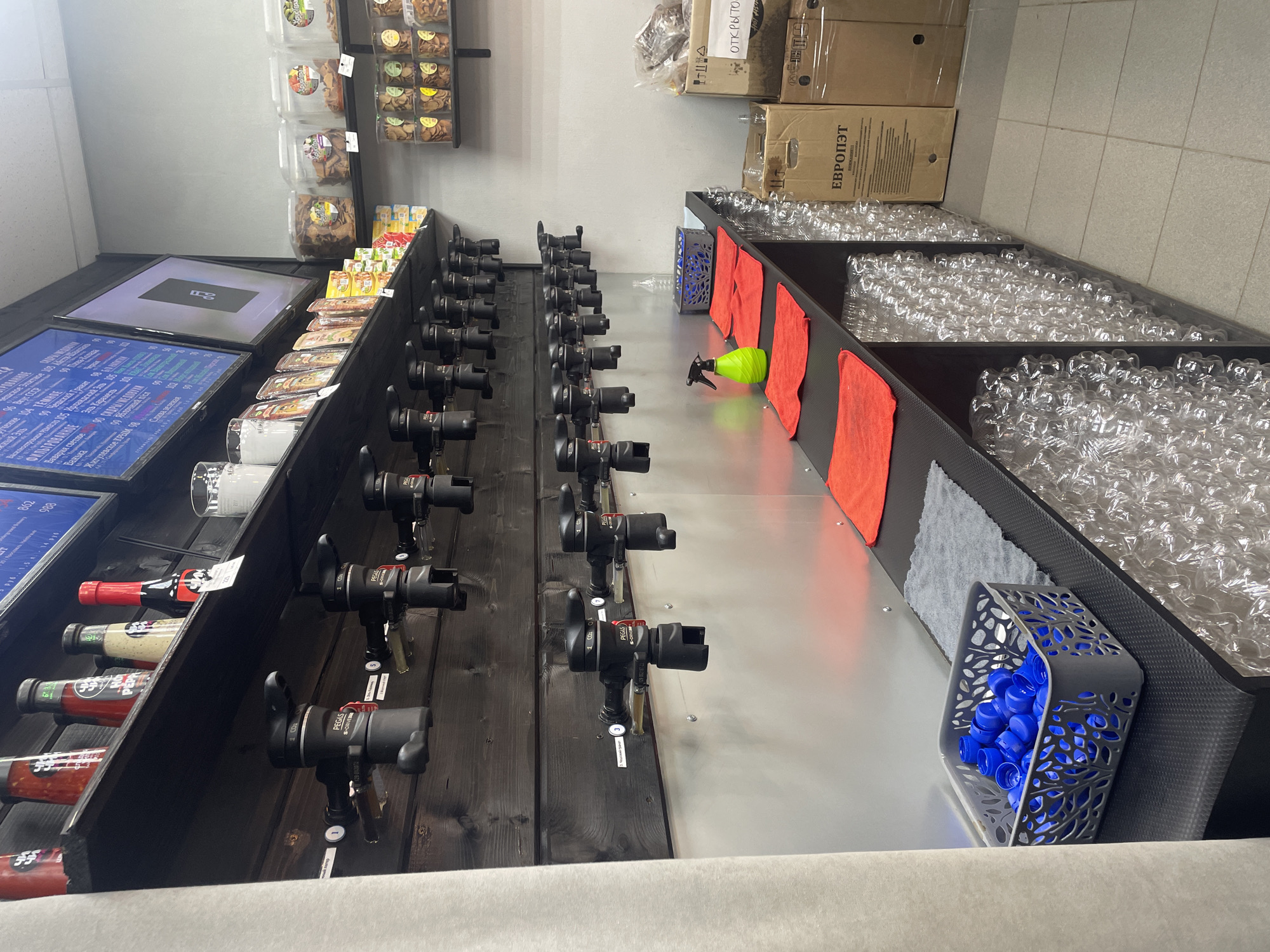 This image shows a retail display for alcoholic and non-alcoholic beverages, likely in a store in Rusanovo, Russia. The main subjects are rows of beer taps, displayed on a dark wooden panel, with various bottles of what appear to be sauces or other condiments in a shelf to the left. Above the bottles and taps, there are shelves stocked with packaged snacks and larger boxes, some with Russian text.
The scene depicts a self-service beverage station. The taps are labeled with prices and product names, such as "Аный Эль," "Вавария светлое," and "Жигулевское КРЫМ," suggesting a selection of beers. There are also clean plastic bottles neatly stacked to the right, ready for customers to fill. A bright green spray bottle and orange cloths are visible near the taps, indicating cleaning supplies are readily available.
The setting appears to be indoors, possibly a small shop or a dedicated section within a larger store. The lighting is bright and even, suggesting it is daytime or the store is well-lit. There are no people visible in the image, focusing the attention entirely on the products and the dispensing system. The overall impression is one of a modern, organized, and customer-friendly retail environment for beverage purchases. Some visible text includes "ЕВРОПЭТ" and "ОТКРЫТО," further confirming the Russian context.