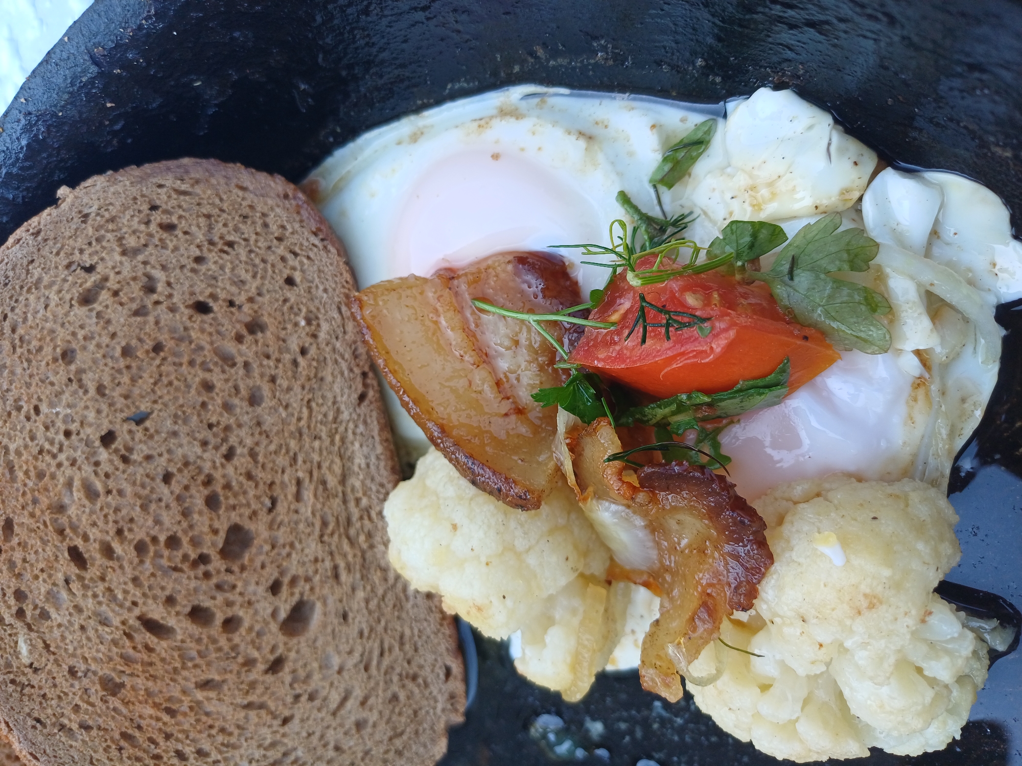 In Sulymivka, Ukraine, a close-up, overhead shot captures a rustic breakfast in a dark cast-iron pan. The main focus is a fried egg, its white spread out and yolk intact, nestled among pieces of cauliflower florets and strips of crispy bacon. A segment of dark, grainy bread is placed to the upper left, partially obscuring the pan's edge. A wedge of tomato and a garnish of fresh green herbs, possibly dill and parsley, add a touch of color to the ensemble. The lighting is natural and appears to be outdoors, with the overall scene suggesting a simple, hearty meal.