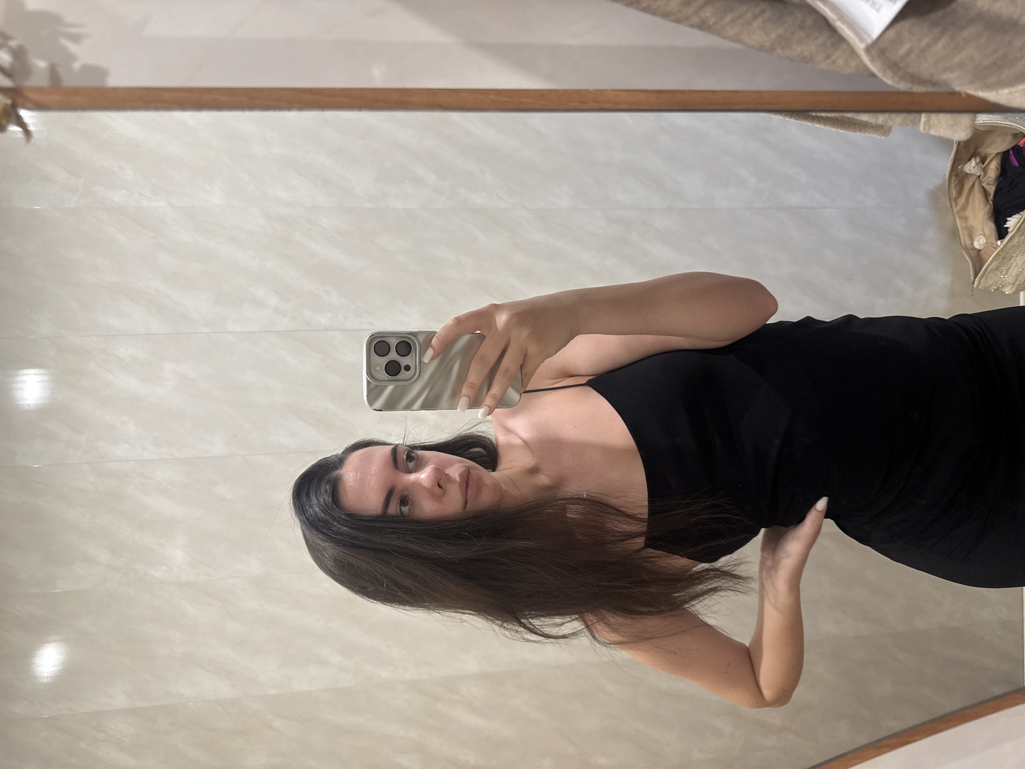 A young woman with long, dark hair is taking a mirror selfie. She is wearing a black, spaghetti-strap dress and has her left hand on her hip. She is holding a silver smartphone in her right hand, which is positioned to capture her reflection. The background appears to be a tiled wall, possibly in a fitting room, with a mirror to her right. A handbag and a beige cardigan are visible in the reflection on the right side of the image.