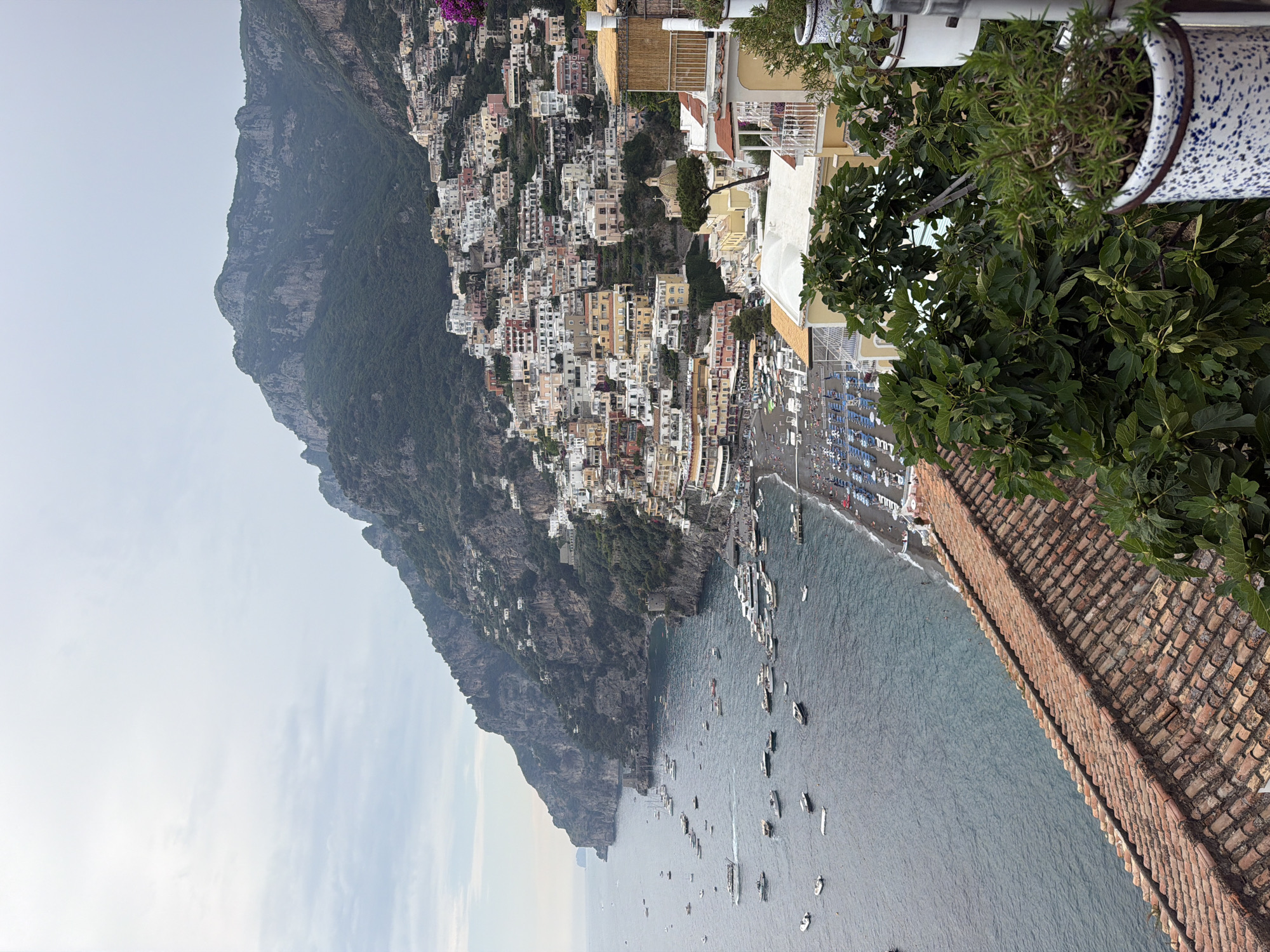 The image captures a high-angle view of Positano, Italy, perched on a steep cliffside. Buildings in various colors and sizes cascade down the slope towards the sea. A rugged, verdant mountain forms the backdrop on the left. The sea is dotted with numerous boats, and a beach with blue sunbeds is visible. A portion of a tiled roof and greenery are in the foreground.
